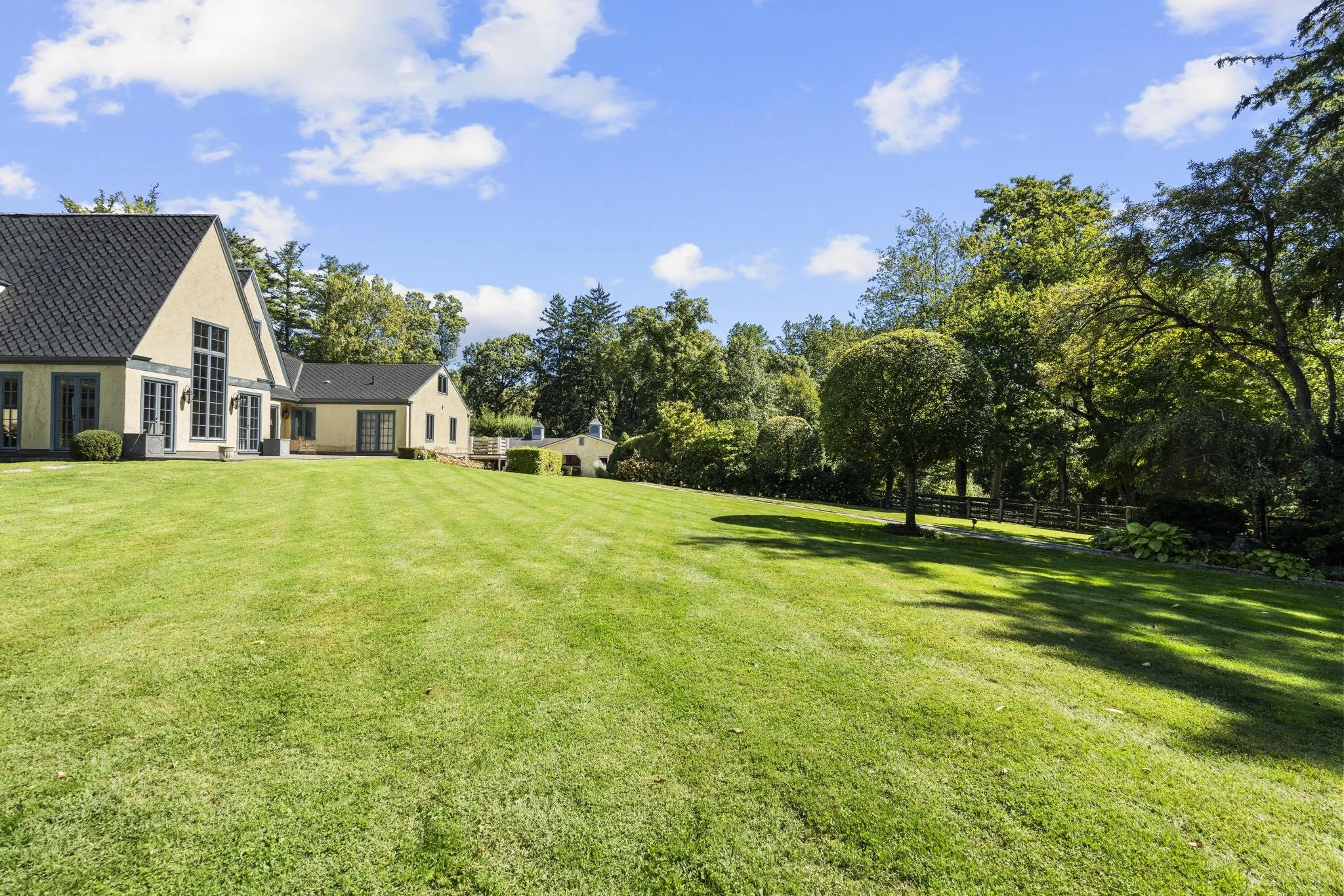 A spacious yard with neatly mowed green grass, bordering trees, and a large house with a black shingle roof and beige exterior walls.