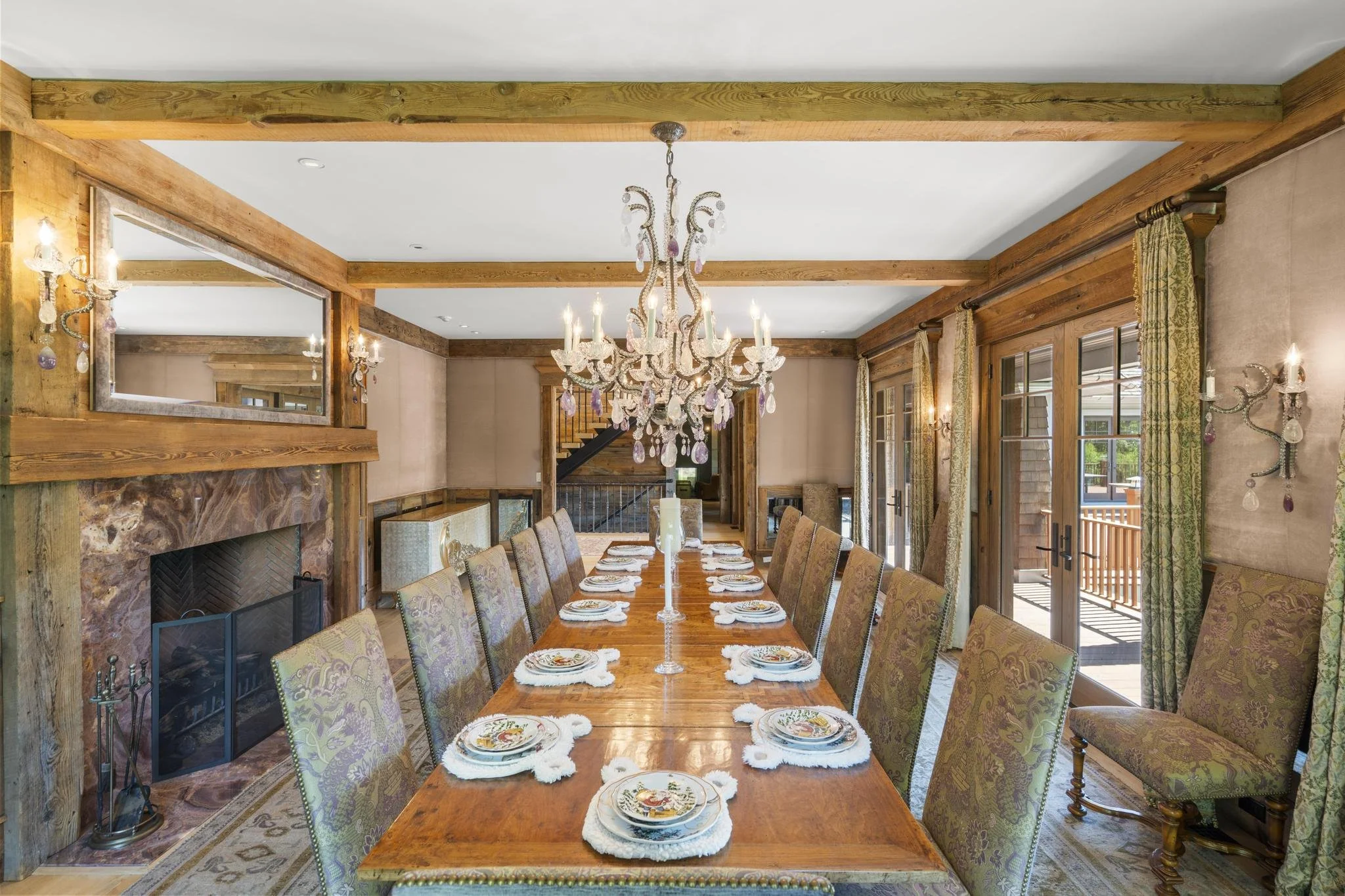 A dining room with a long wooden table set with plates and napkins. An ornate chandelier hangs above the table, and there are windows with curtains and a sliding door leading outside. The room features wood beams and a fireplace with a mirror above it.