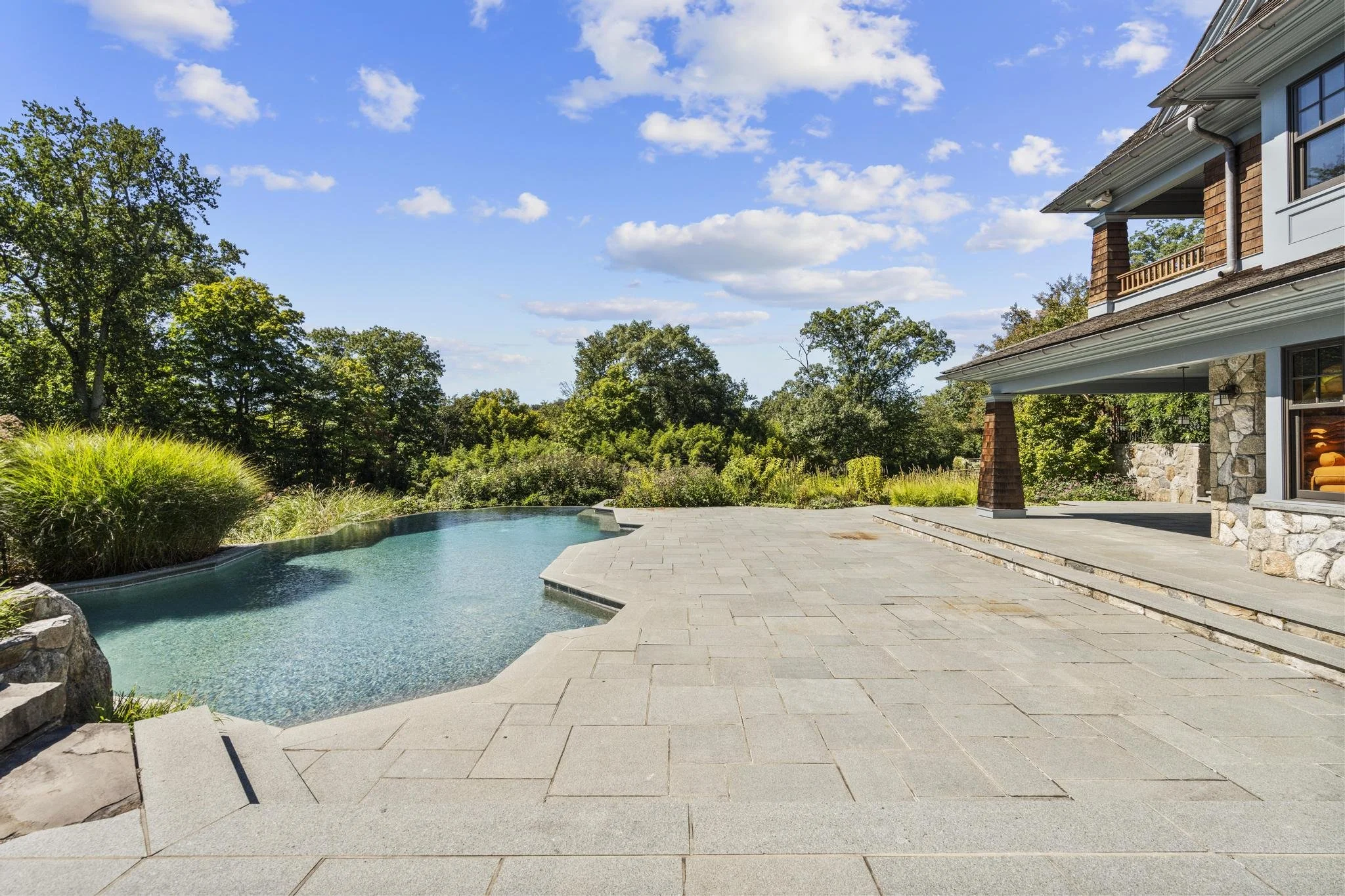 Backyard patio with a swimming pool, gray paving stones, lush green landscaping, and a two-story house with a partial view of the roof and windows, under a bright blue sky with white clouds.