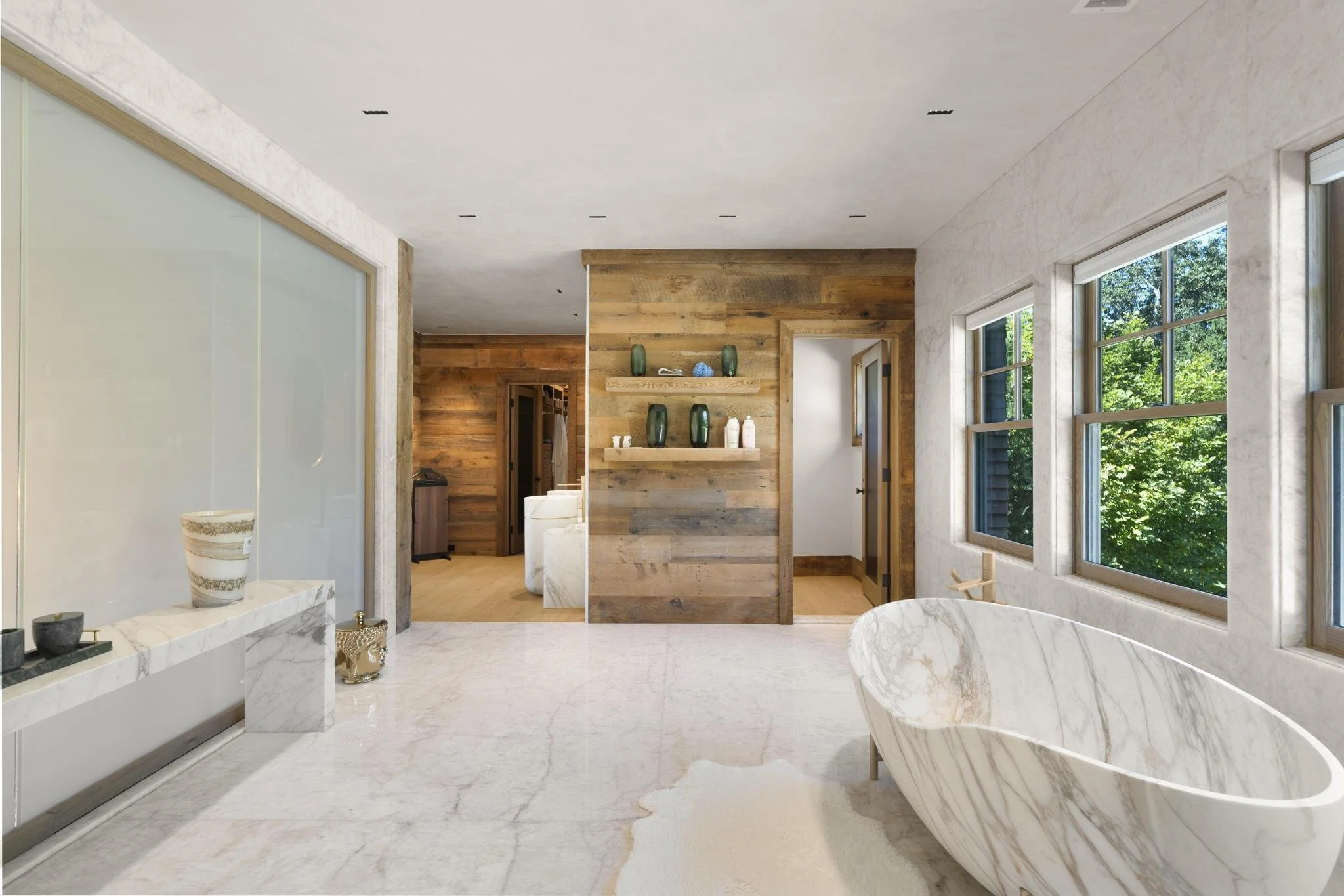 Modern bathroom with marble bathtub, marble flooring, large windows, and a wooden partition with decorative vases.