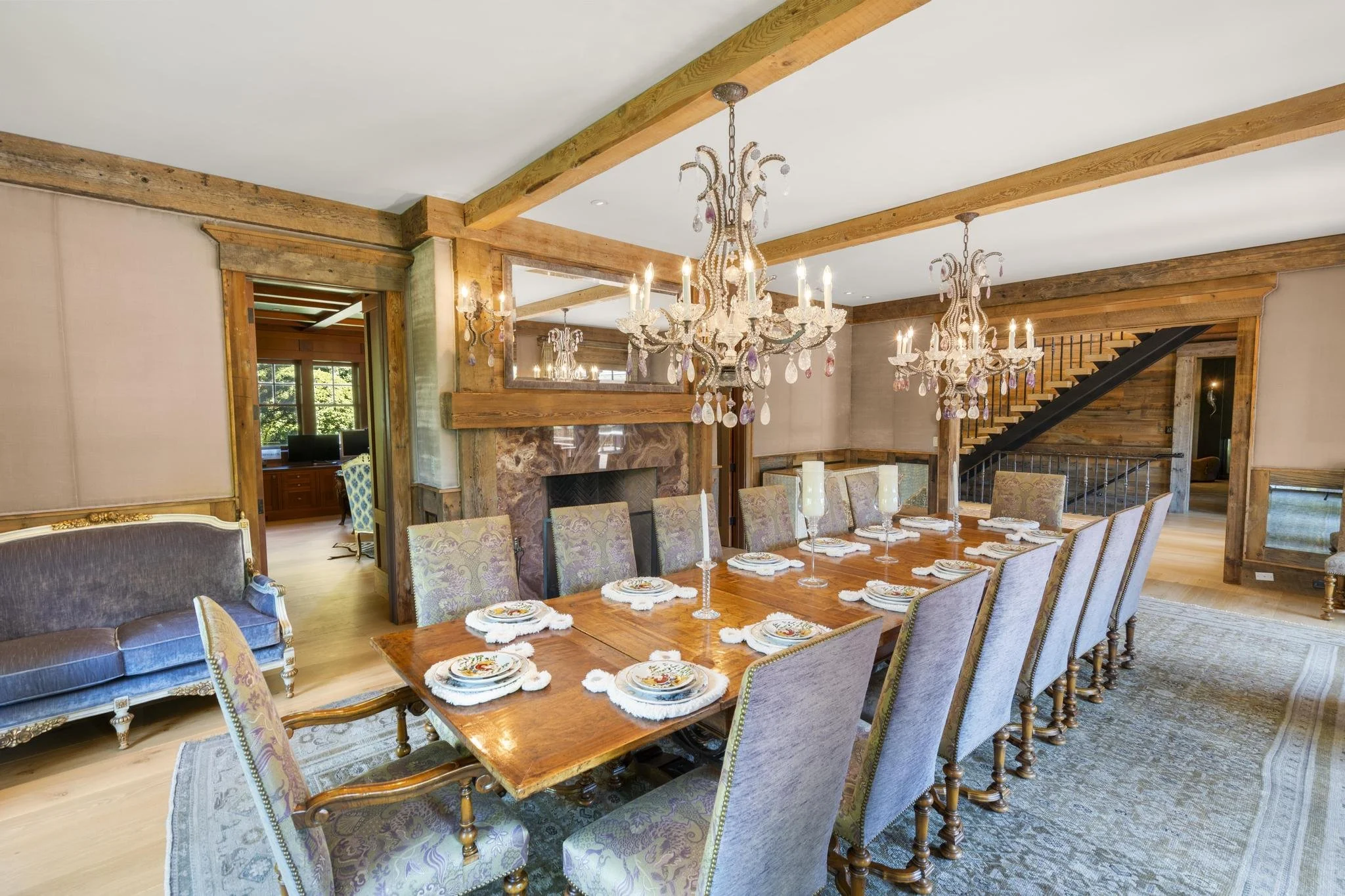 A dining room with a long wooden table set with plates, silverware, and candles, surrounded by upholstered chairs, and two ornate chandeliers hanging from a ceiling with exposed wooden beams.