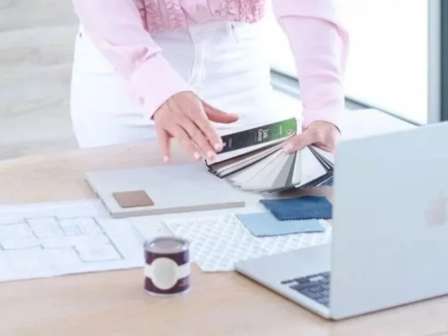 Person holding and flipping through a color swatch book at a desk with design samples, a laptop, and architectural plans.