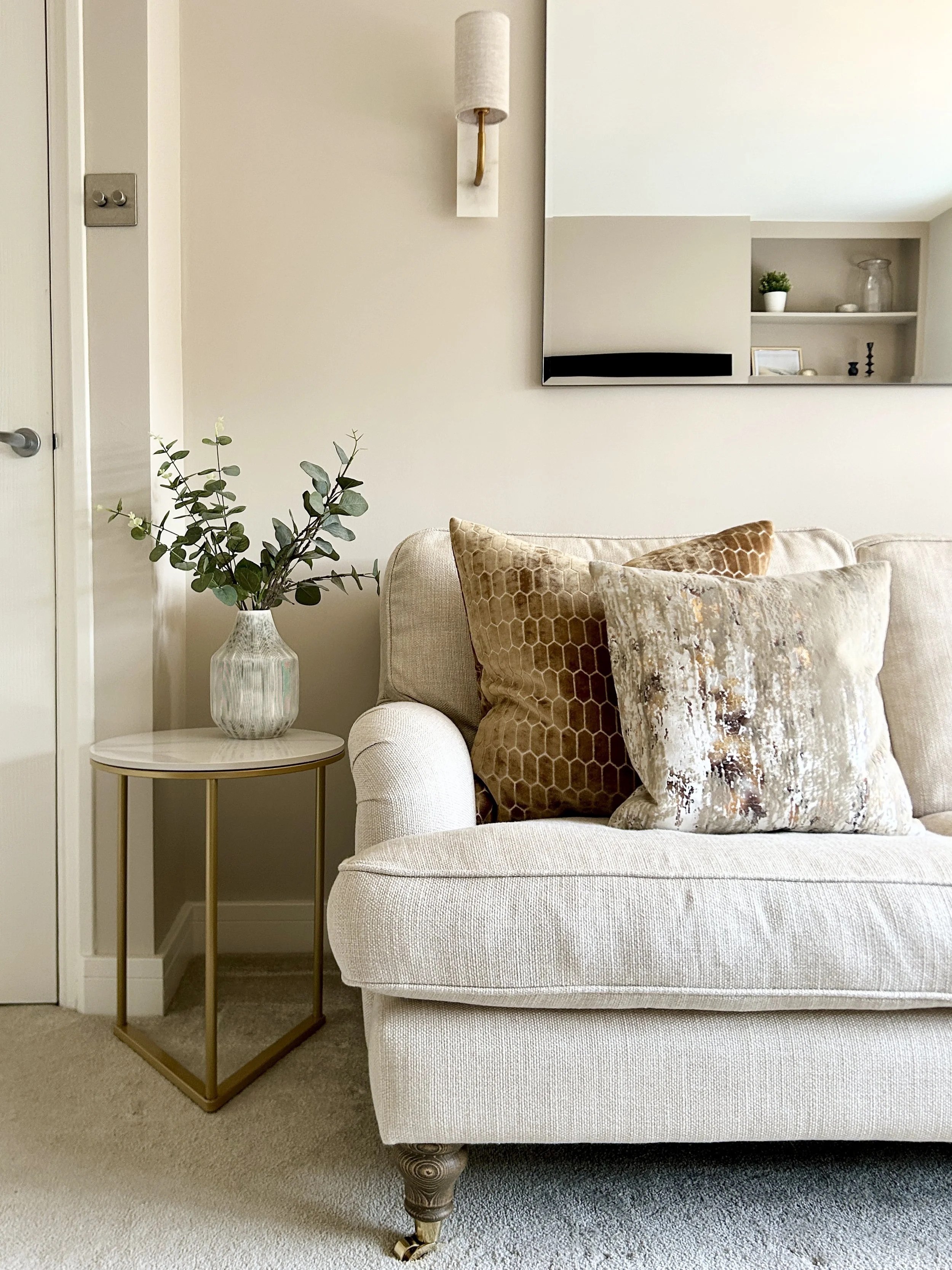 Living room corner with white upholstered sofa, decorative pillows, glass-topped side table with gold frame and a vase of greenery, and wall mirrors with shelves and decorative items in the background.