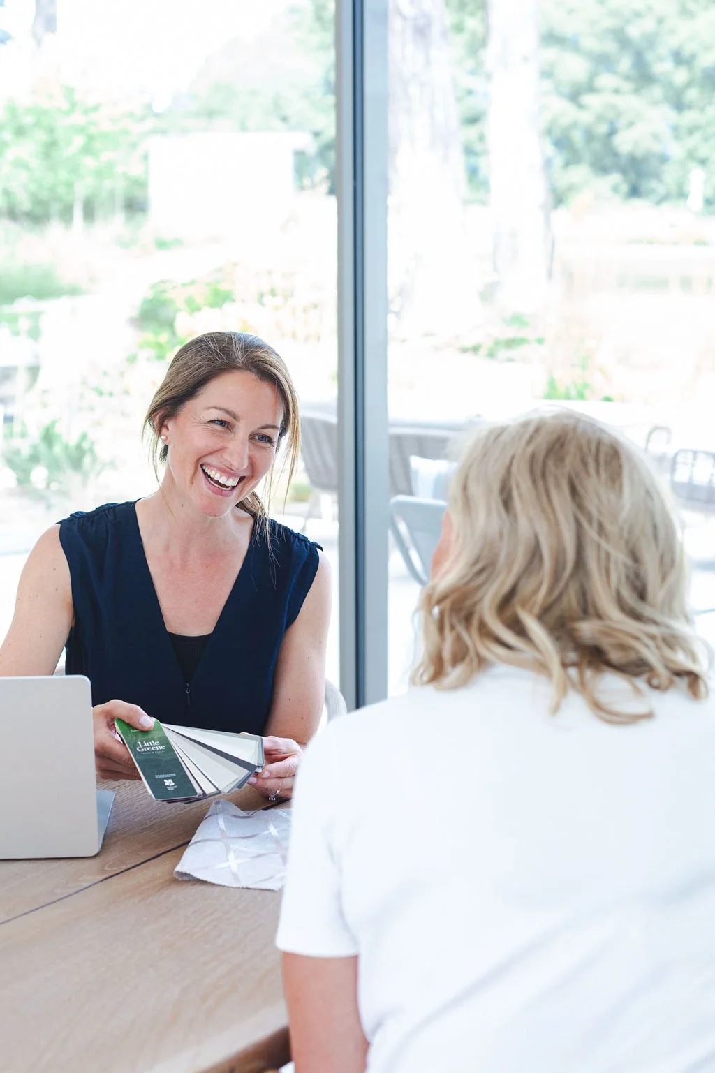 A woman showing color samples to another woman in a bright indoor setting with large windows and a garden view outside.