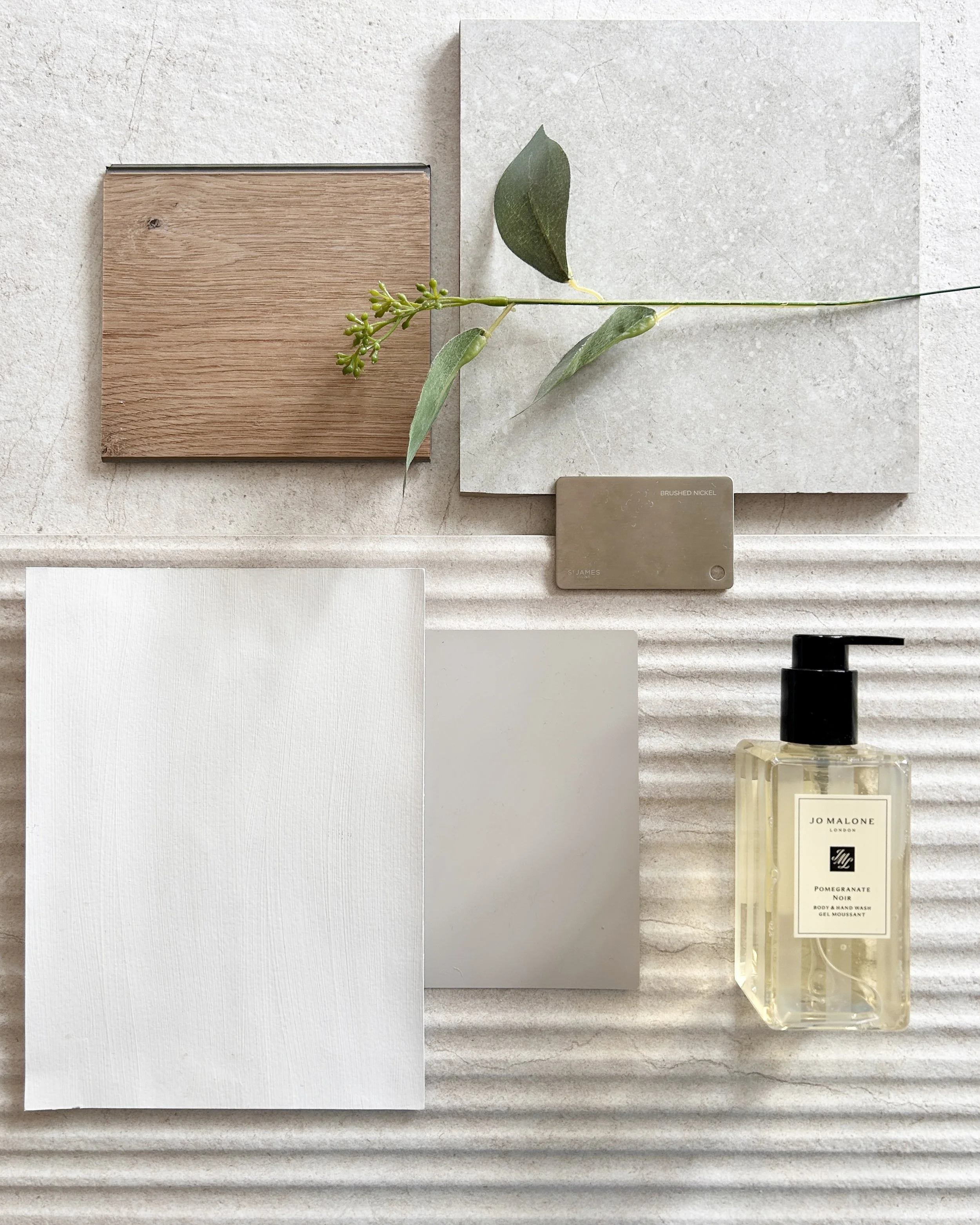 A flatlay of various materials including two ceramic tiles, a piece of wood, a color sample card, a silver paint swatch, a sprig of eucalyptus, and a bottle of Jo Malone Pomegranate Noir perfume.