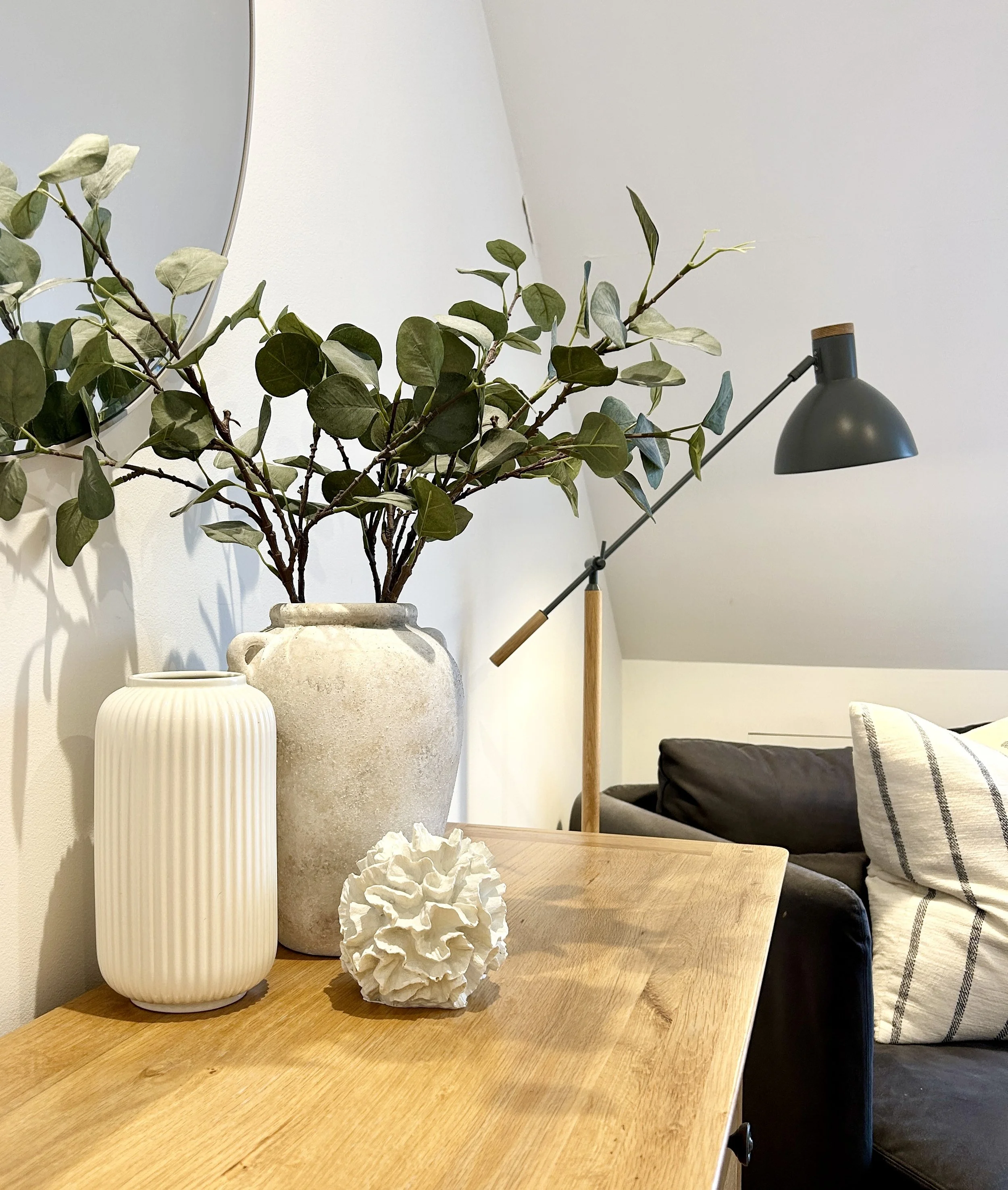 A wooden table with a tall white ceramic vase holding green leafy branches, a large textured beige vase, a crumpled white paper sculpture, and a small white container. In the background, there is a black wall lamp and a dark sofa with striped pillows, a mirror on the wall, and plain white walls.