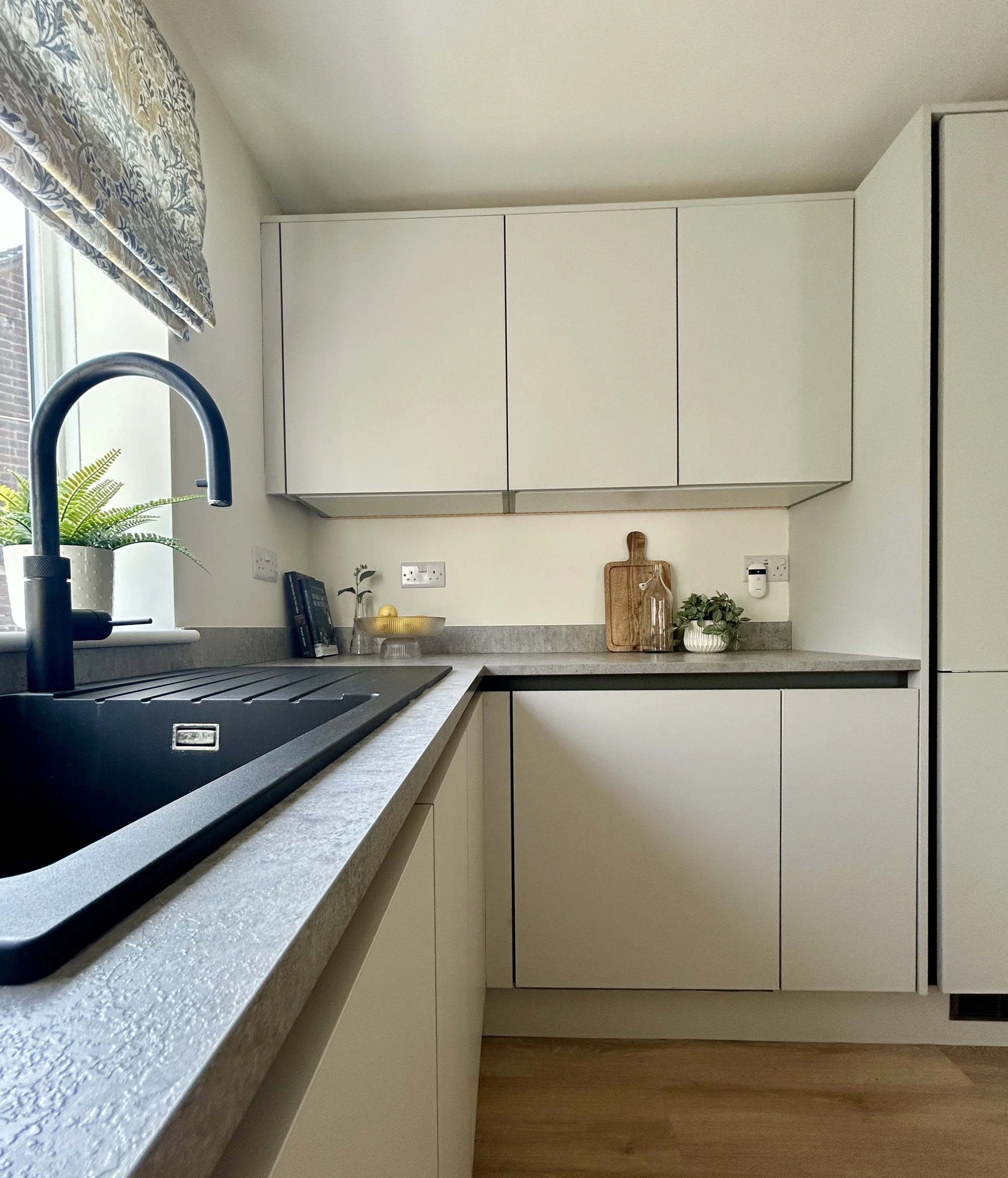 A modern kitchen with a gray speckled countertop, a black sink with a curved faucet, white cabinets, a window with floral curtains, and small decorative items on the counter including a potted plant, a fruit bowl, and a wooden cutting board.