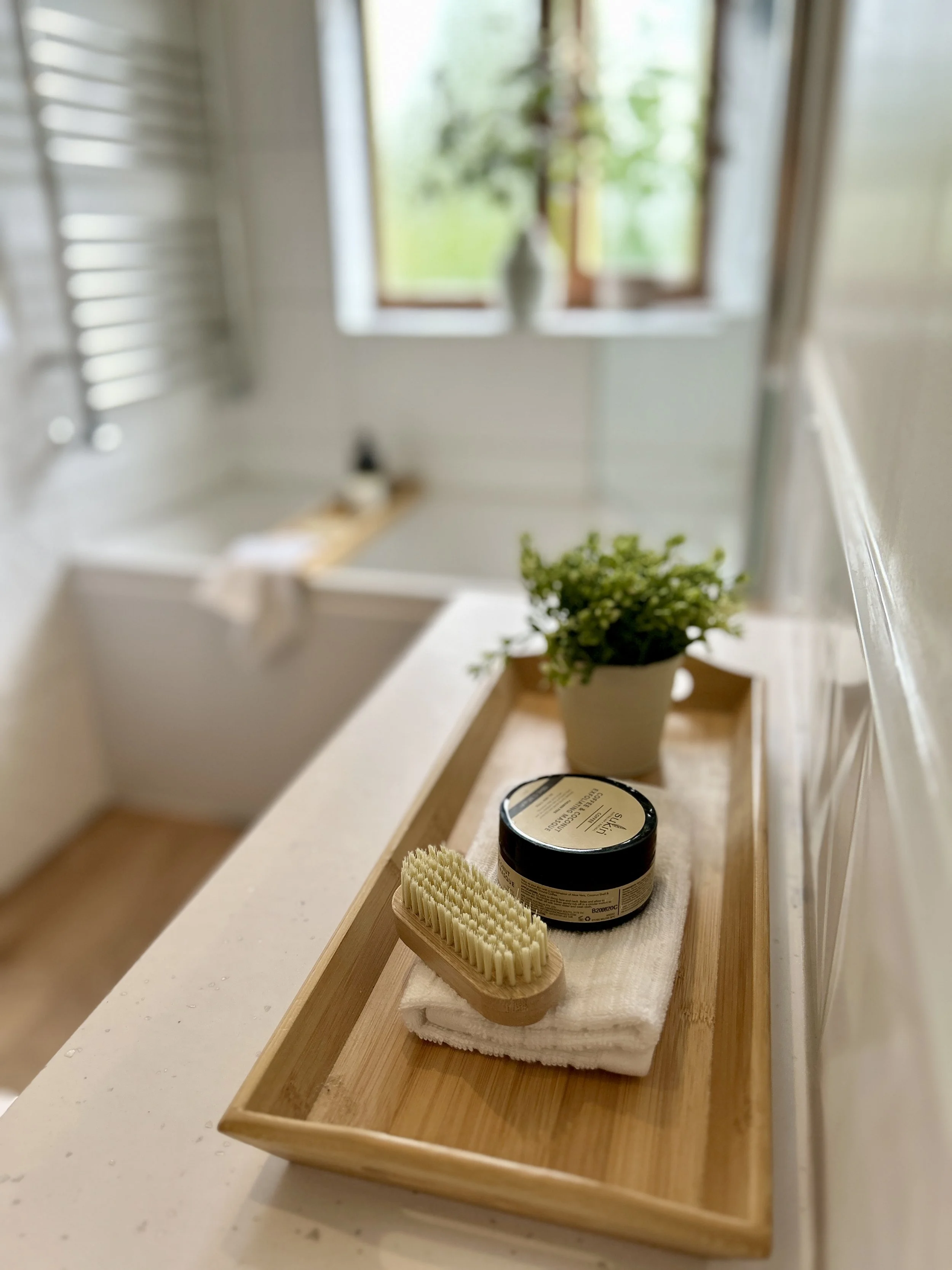 A wooden tray on a countertop with a jar of hair styling product, a hairbrush, and a folded white towel, with a green plant and a window in the background.