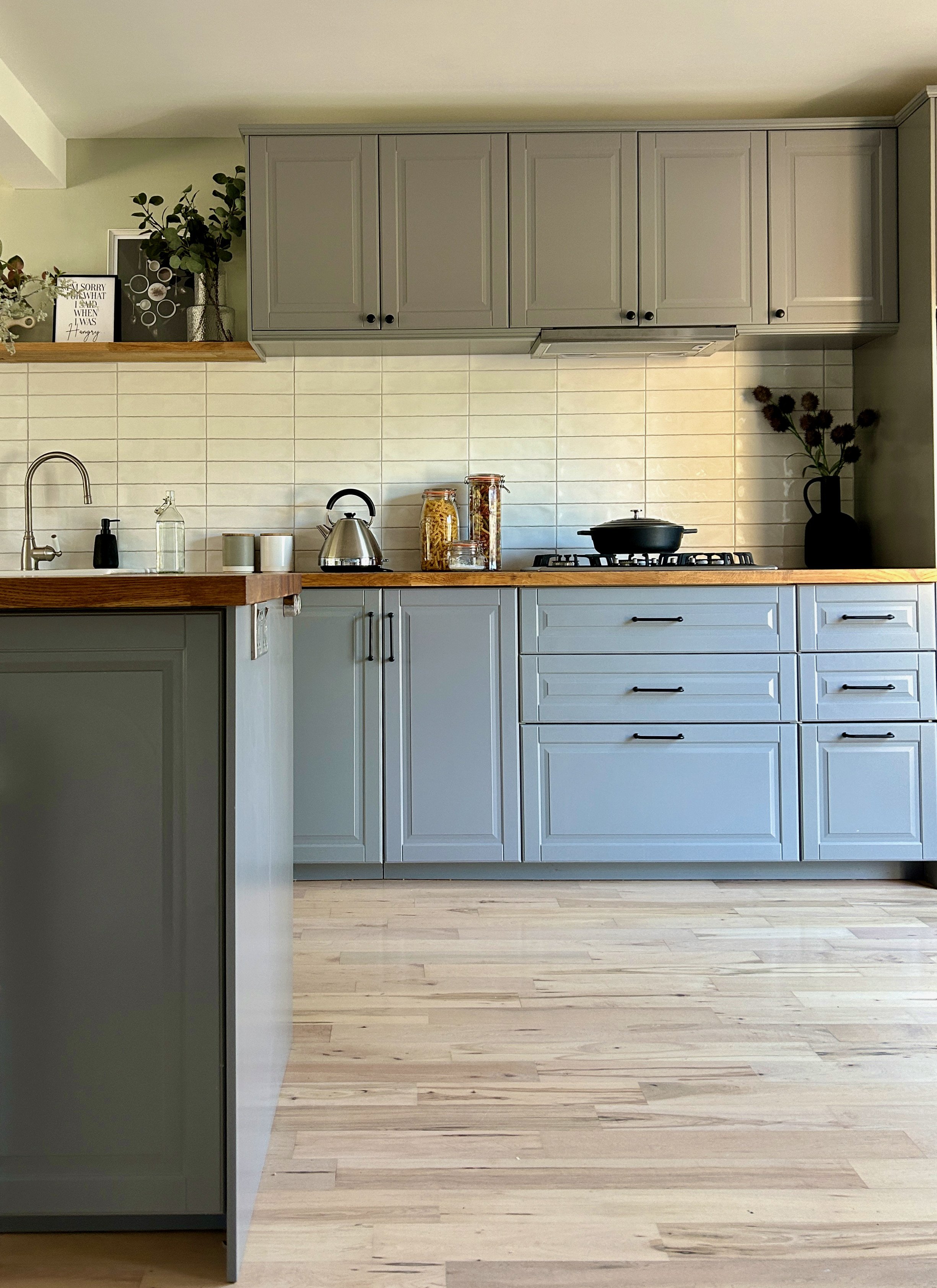 A modern kitchen with light blue cabinets, a wooden countertop, and a light wood floor. Decor includes jars with pasta, a black stove, a kettler, a faucet, and plants on top of the cabinets.