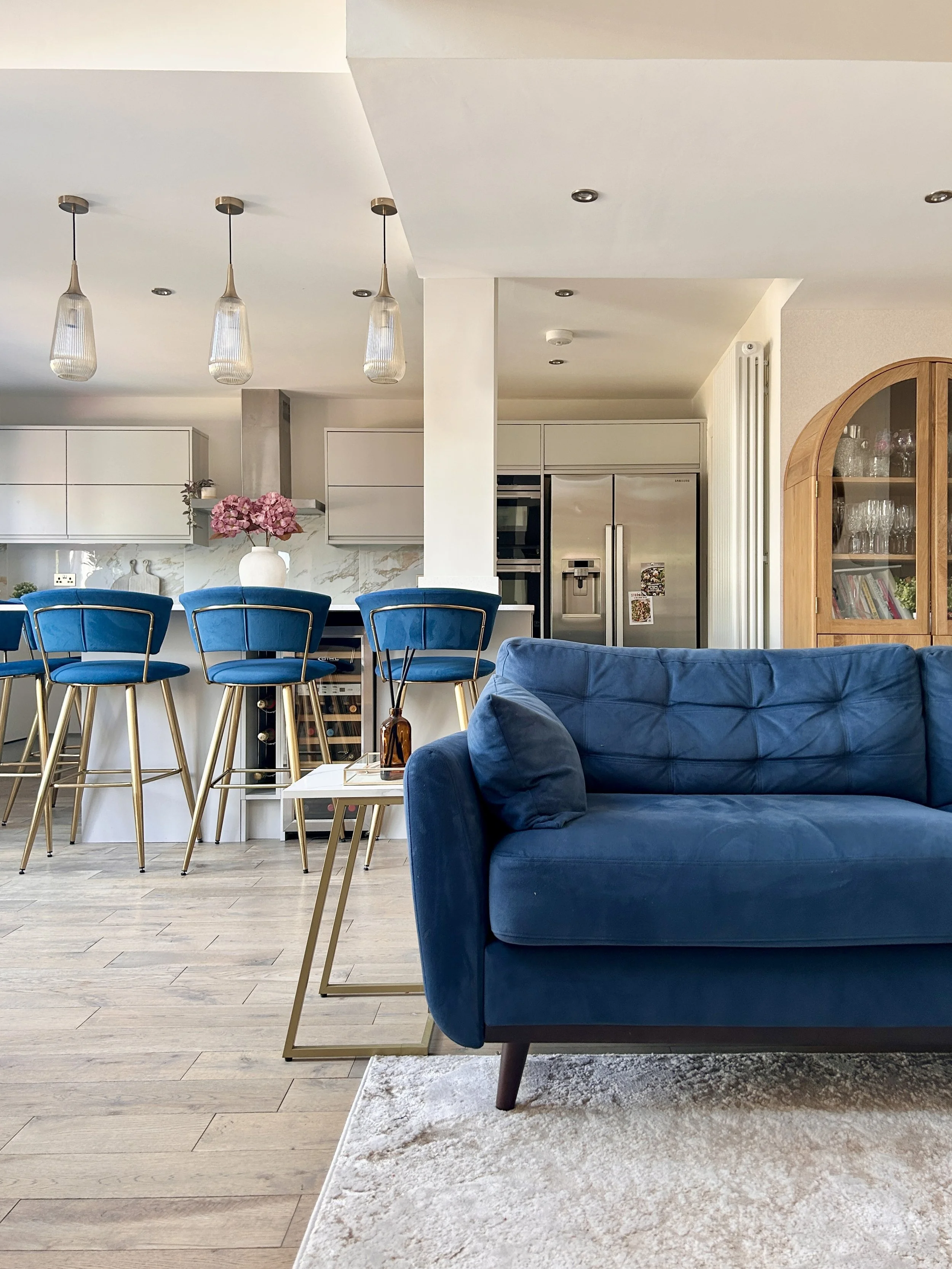 Modern living room and kitchen with blue velvet sofa, high-backed blue chairs at the kitchen counter, white cabinets, and decorative pink flowers in a white vase.