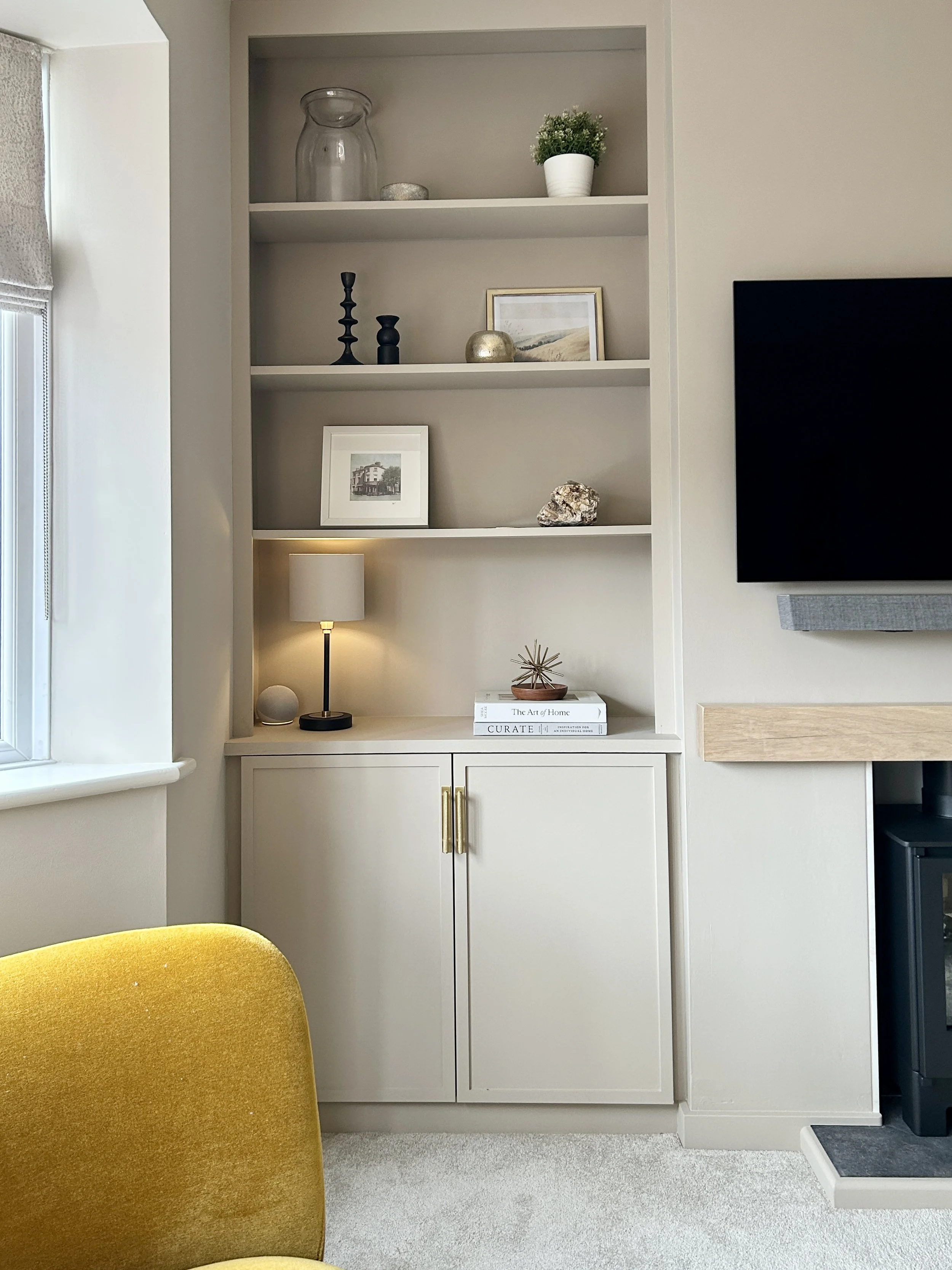 Living room with white built-in shelves containing decorative items like vases, picture frames, and a small potted plant. There is a black TV mounted on the wall to the right, next to a light wood fireplace mantel. A yellow chair is partially visible in the foreground, and a window is on the left side of the image.
