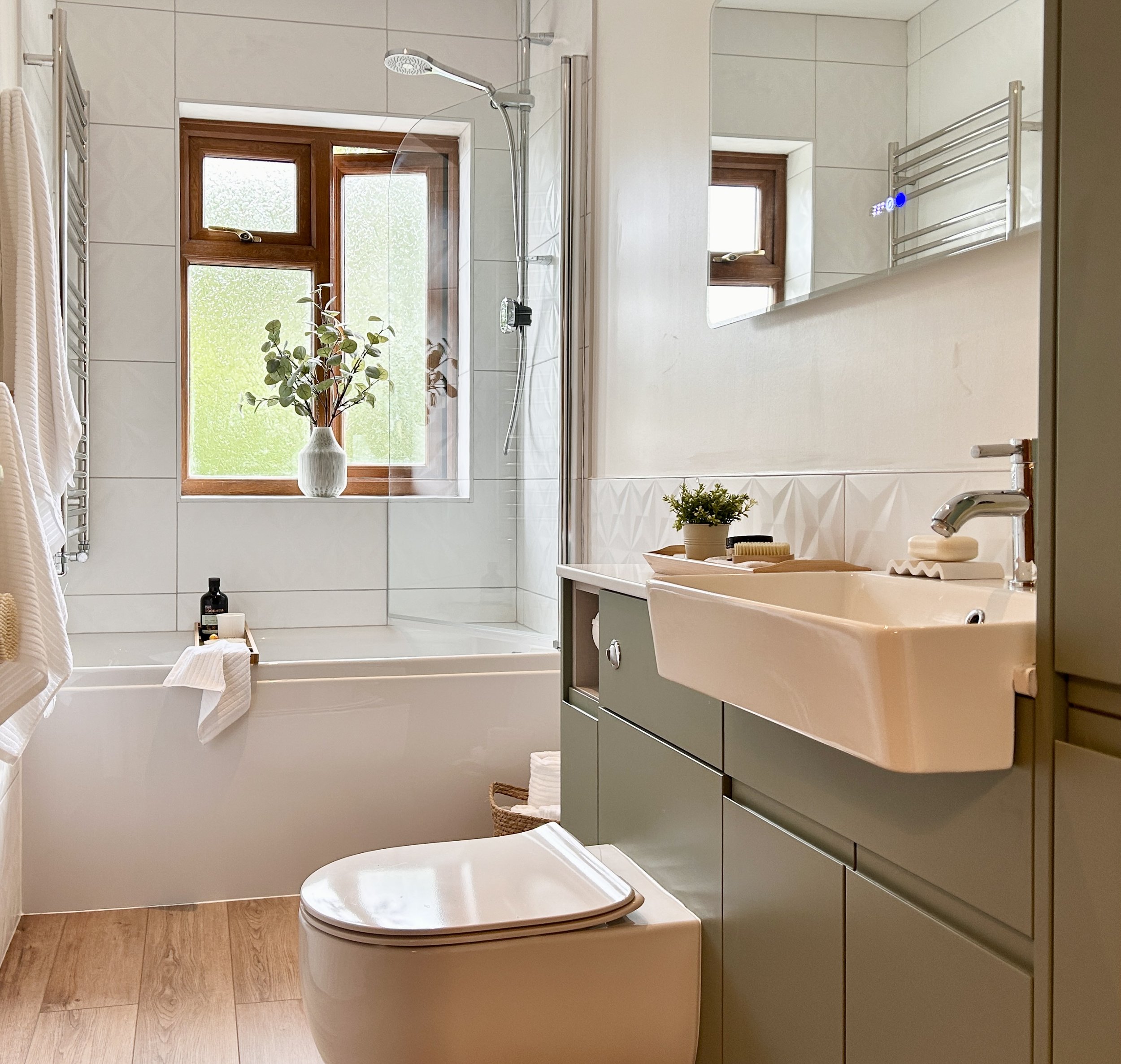 A cozy bathroom with a bathtub and a walk-in shower, decorated with wooden frames around the windows, a potted plant on the bathtub, and a vanity with toiletries and a plant. Natural light streams through the windows, creating a warm and inviting atmosphere.