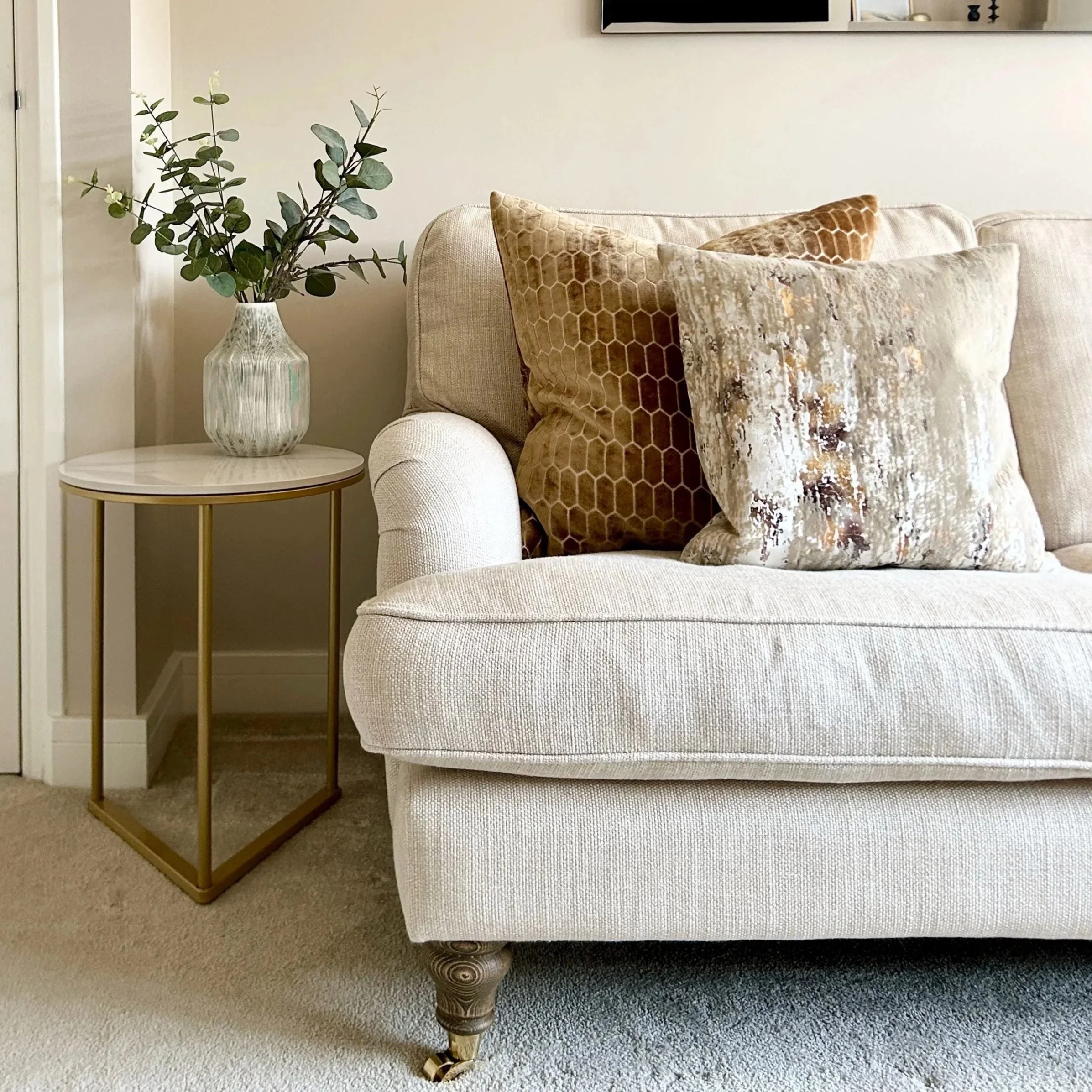 A beige sofa with decorative pillows, next to a small round side table with a floral patterned vase holding green leafy branches, in a living room with beige carpet and a wall-mounted TV.
