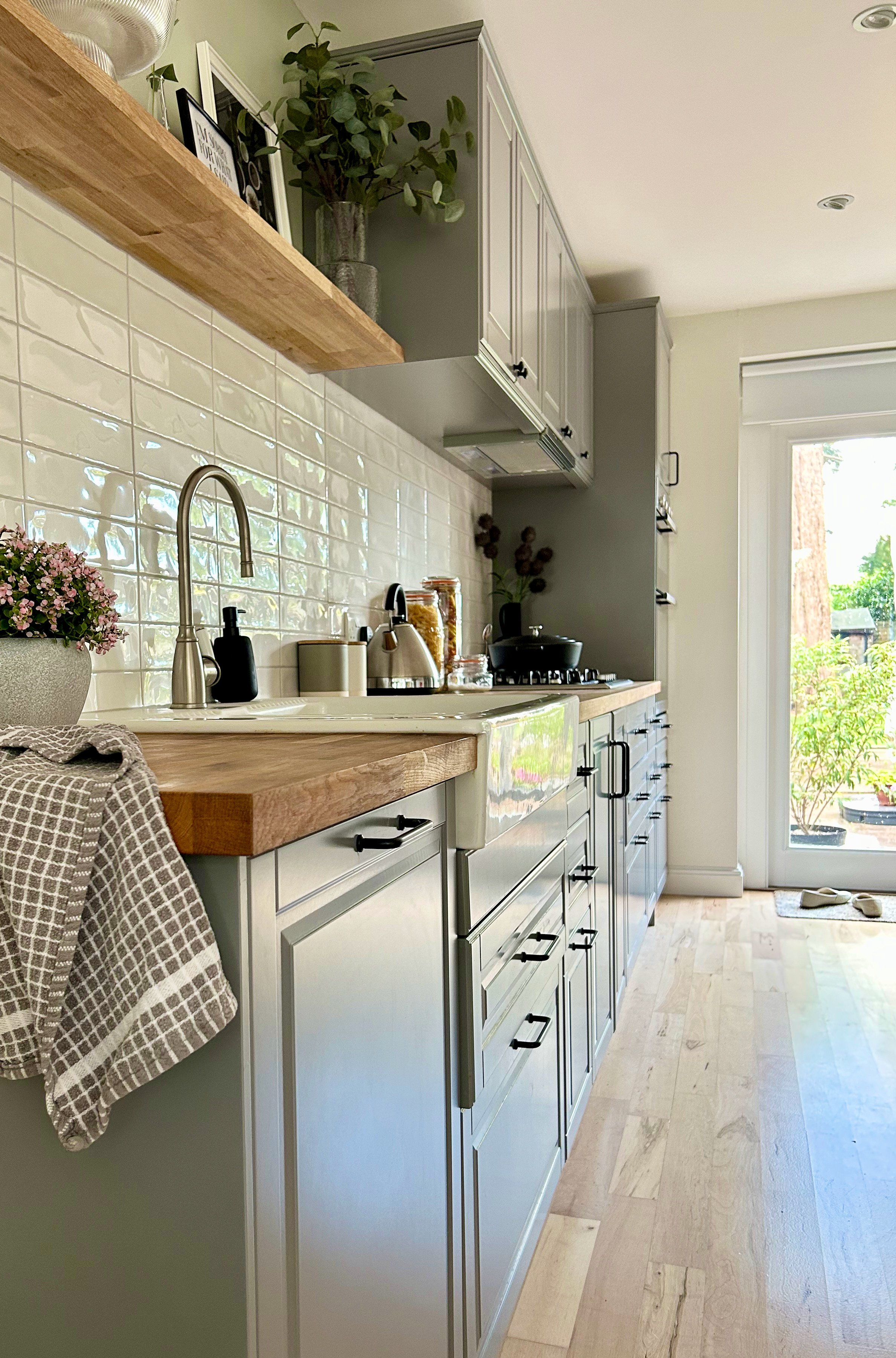 A modern kitchen with white cabinets, wooden countertop, and a beige tiled backsplash. The countertop holds a potted pink flower, soap dispenser, and kitchen utensils. There are glass jars with pasta and other items, a black kettle, and a small black cooking pot on the stove. Above, there are gray cabinets, and open shelving with décor and greenery. A door with a glass panel opens to an outside garden area with trees and plants.