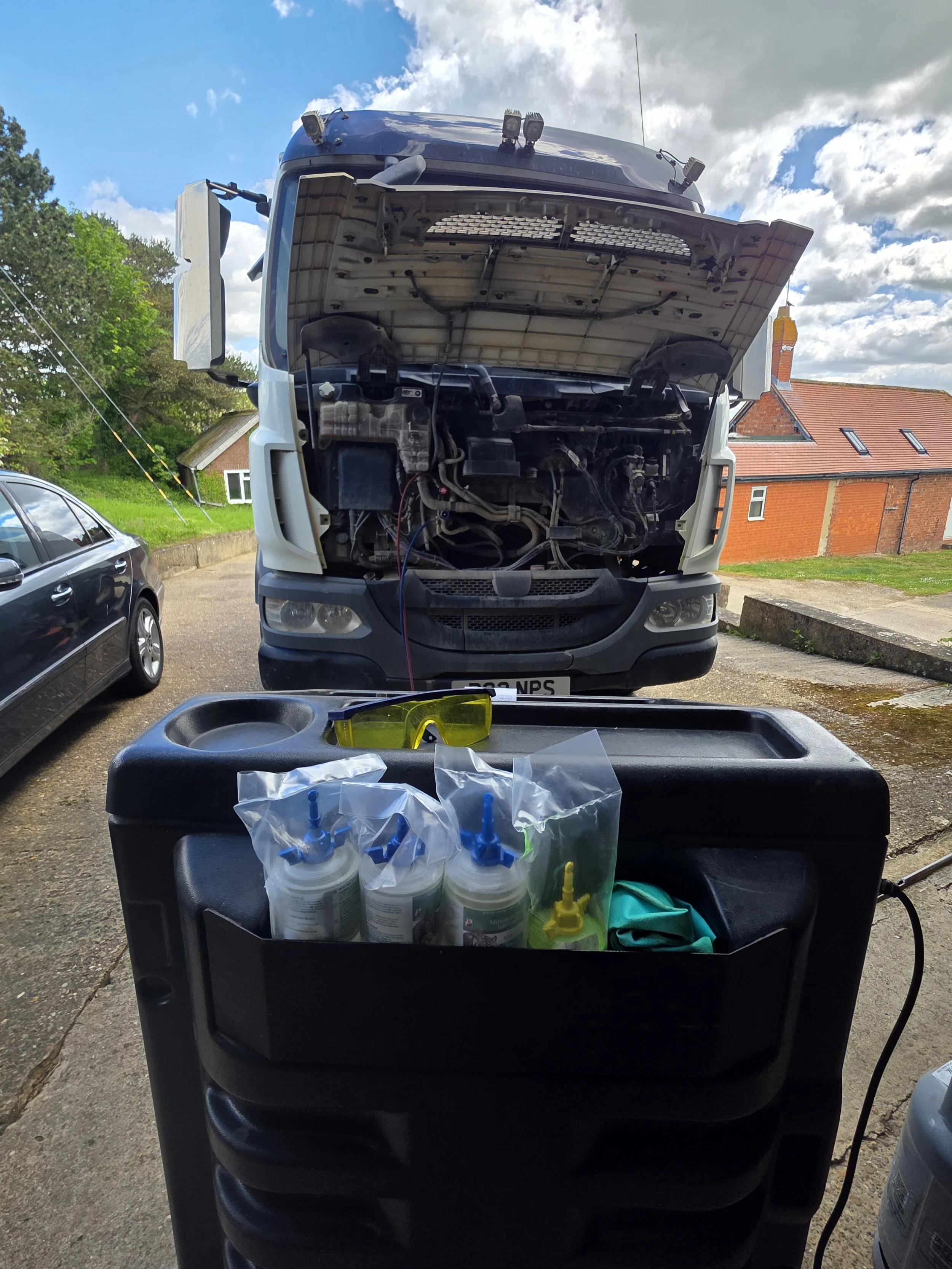 A large truck with its hood open, repairs being carried out, with various tools and supplies in the foreground, and a house with a red roof and cloudy sky in the background. Road sweeper air conditioning service. NPS service and hire