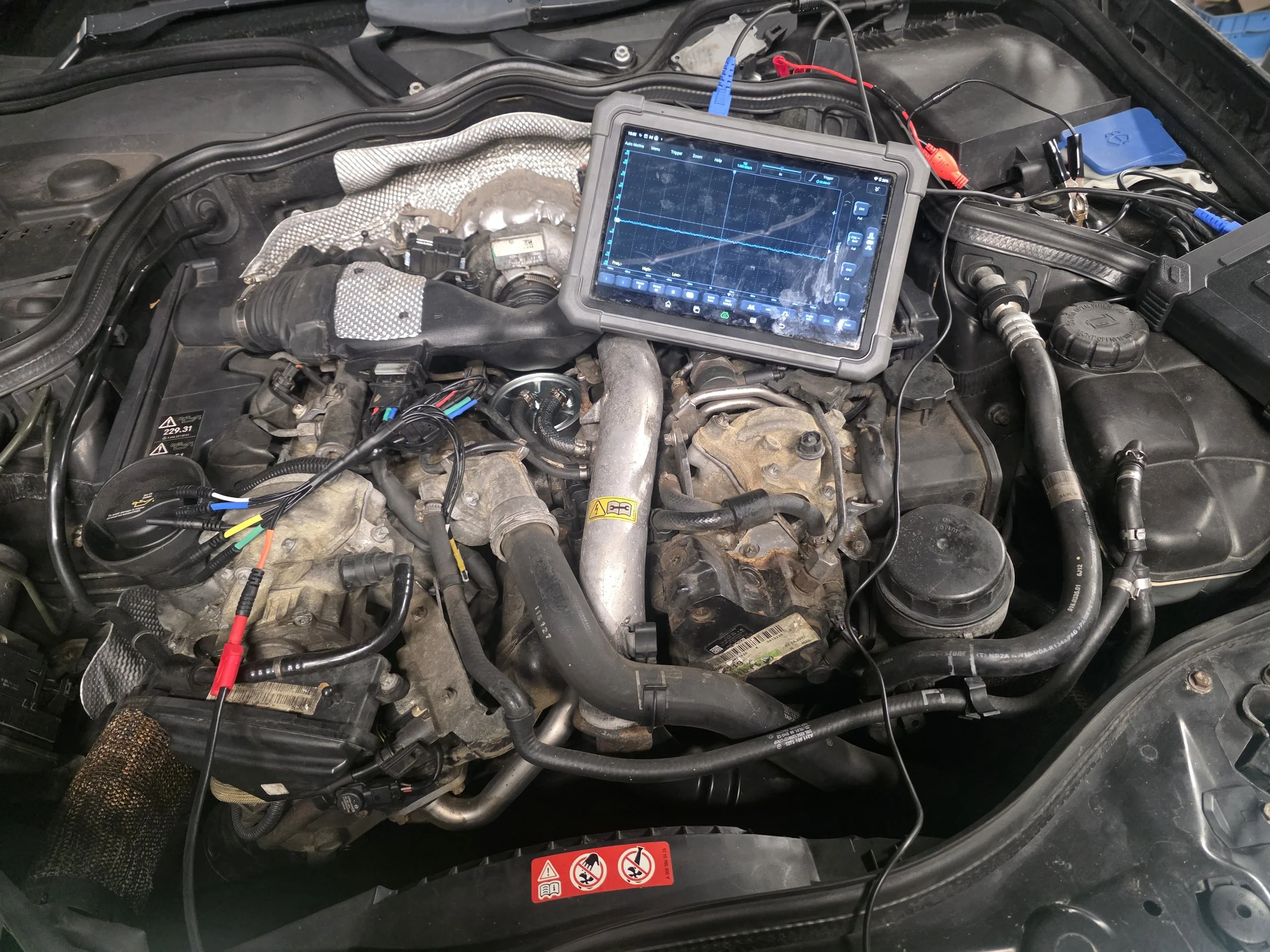 Under the hood of a vehicle, with diagnostic equipment connected to the engine, showing a screen with waveforms and data readings.