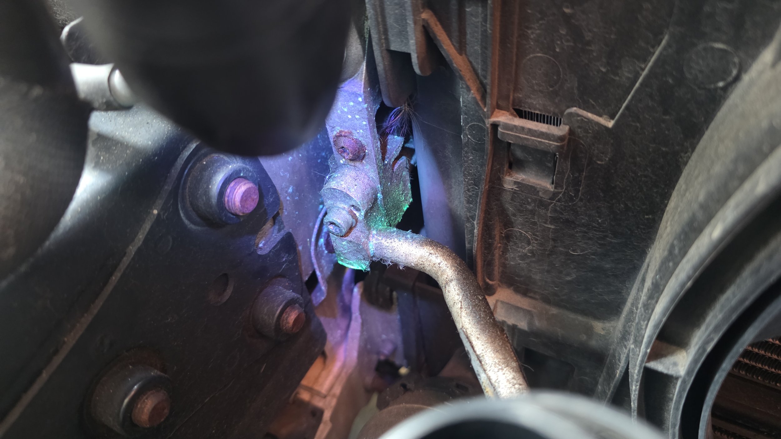 Close-up of an engine component with metal parts, some bolts, and tubes. There are signs of rust and dirt, with lighting revealing some metallic and potentially welded surfaces. Air conditioning service and leak diagnostic using Ultra violet dye UV