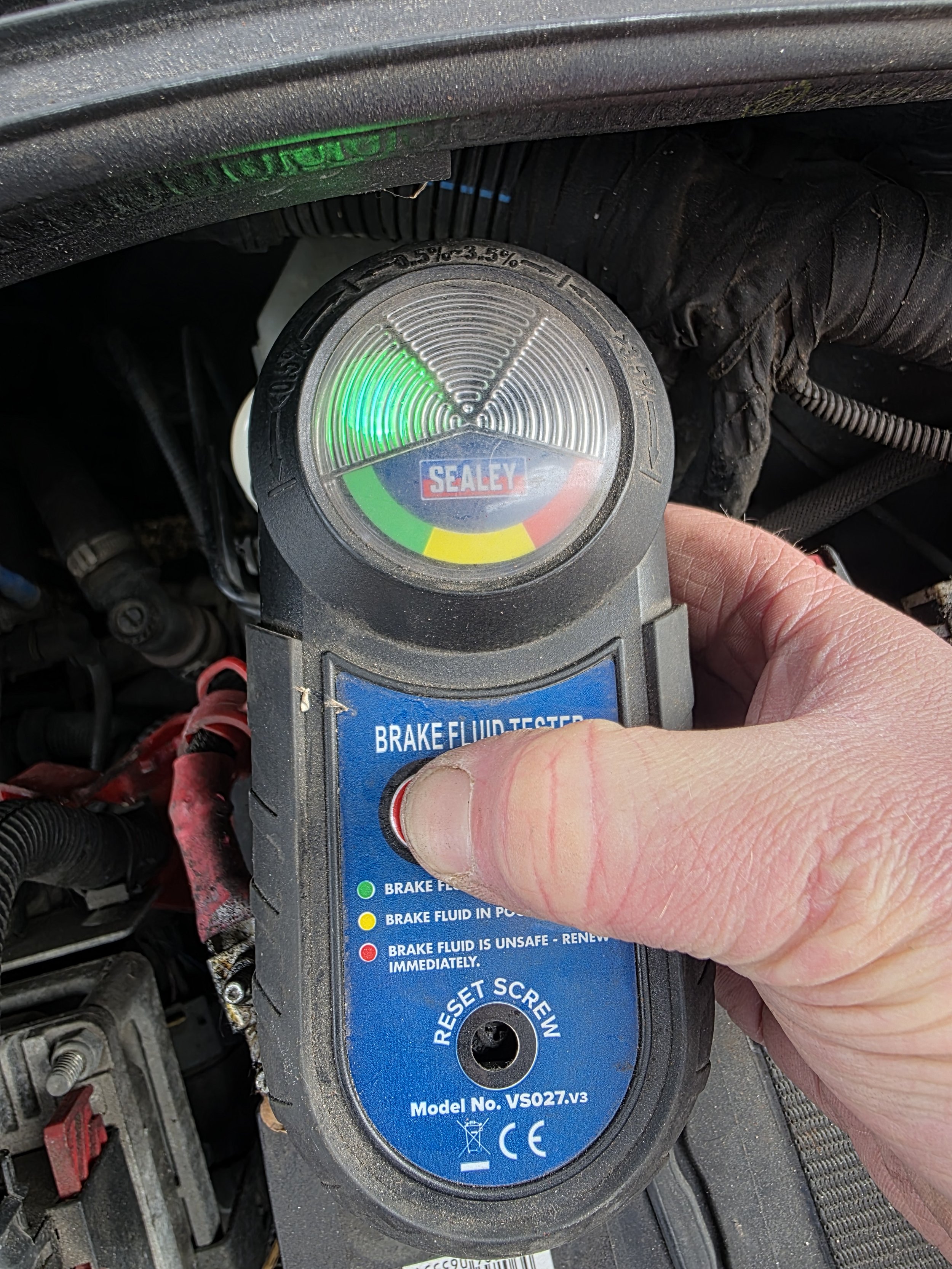 Person holding a blue brake fluid tester near a vehicle's engine, with various wires and engine components visible in the background. Brake fluid test passed
