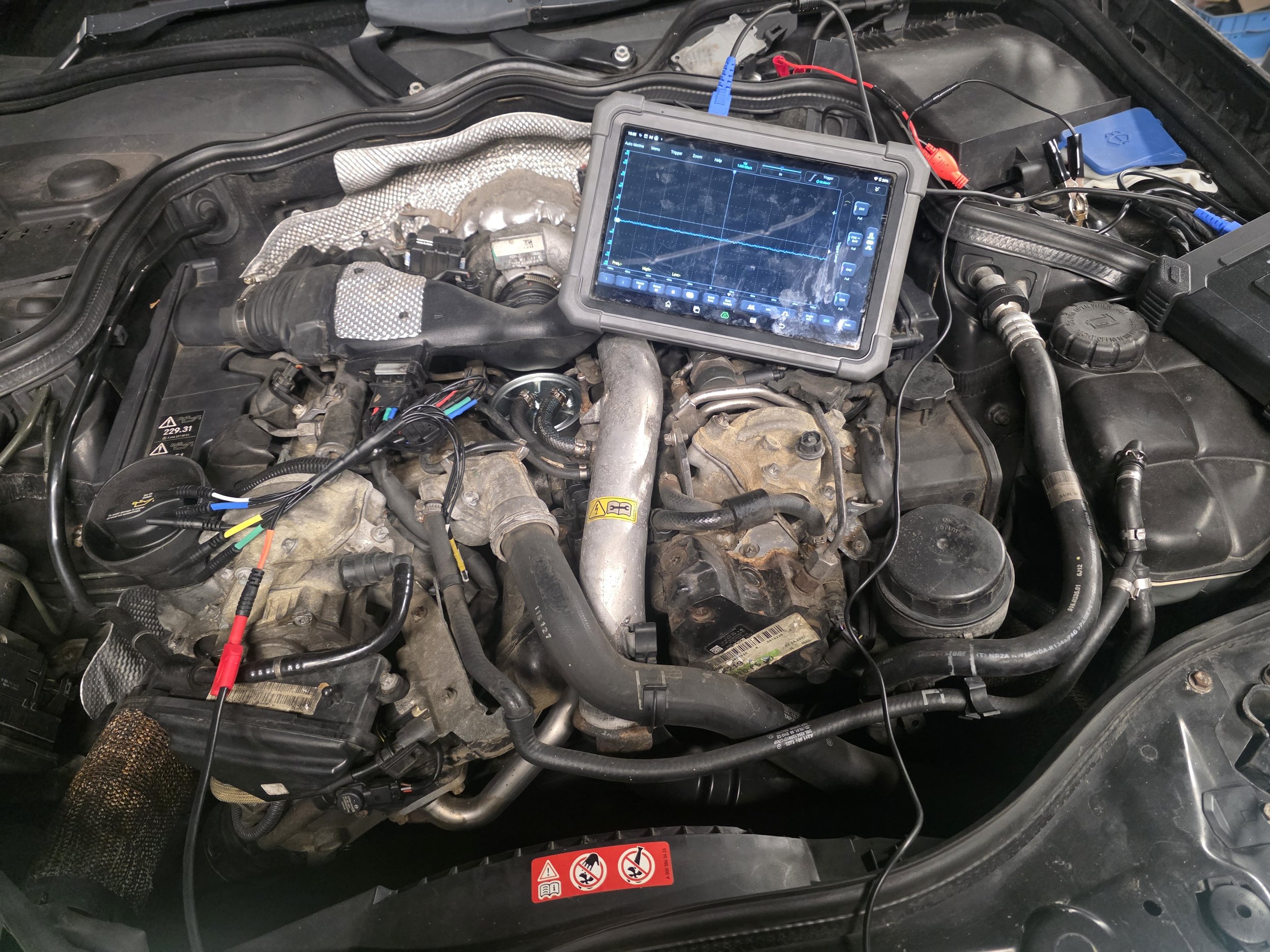 A vehicle engine with diagnostic equipment connected, displaying a graph on a screen placed over the engine bay. Launch oscilloscope in action diagnosing a mass air flow fault