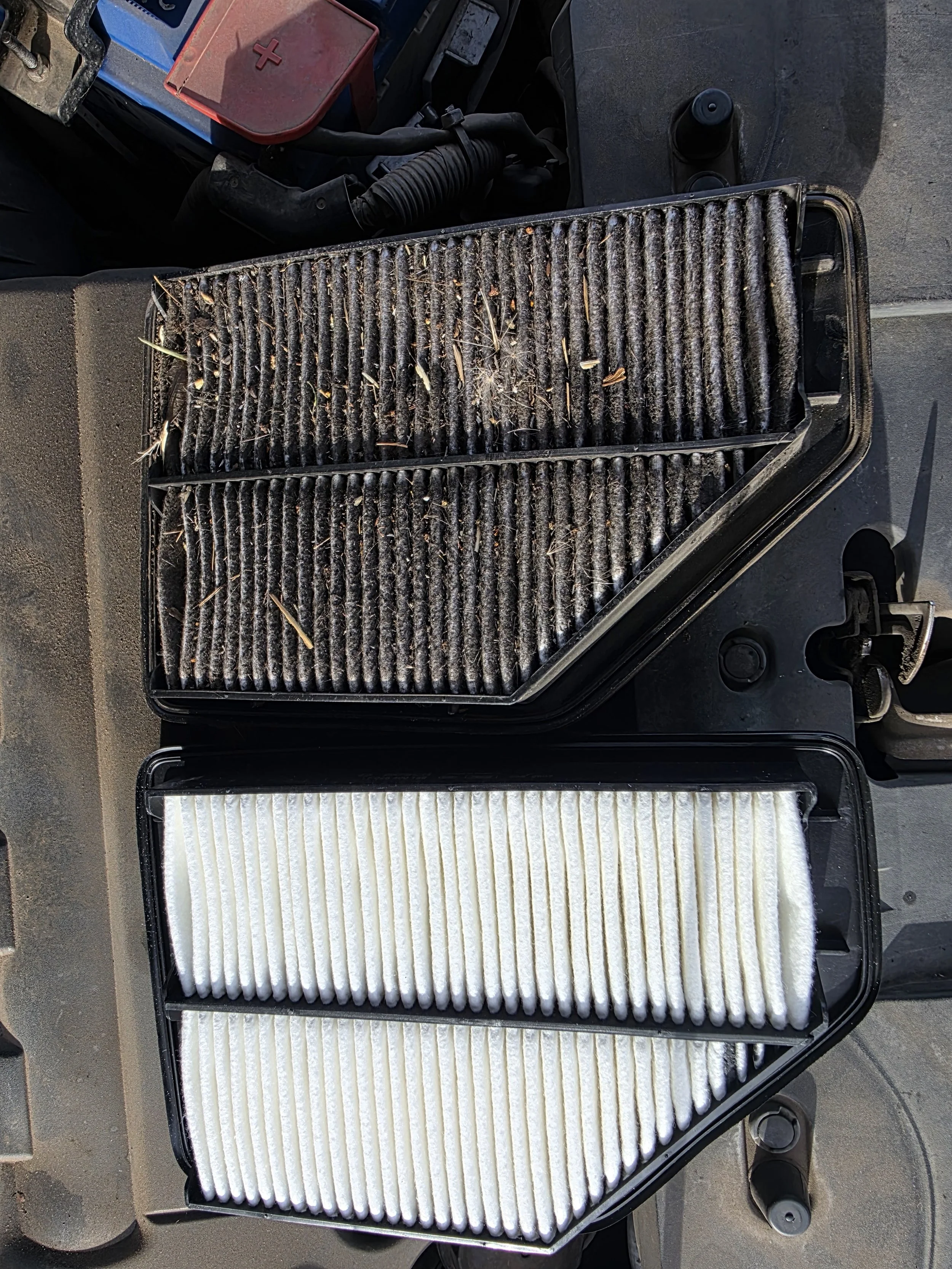 A dirty vehicle air filter and a clean white air filter in black plastic cases, placed on a grey surface.