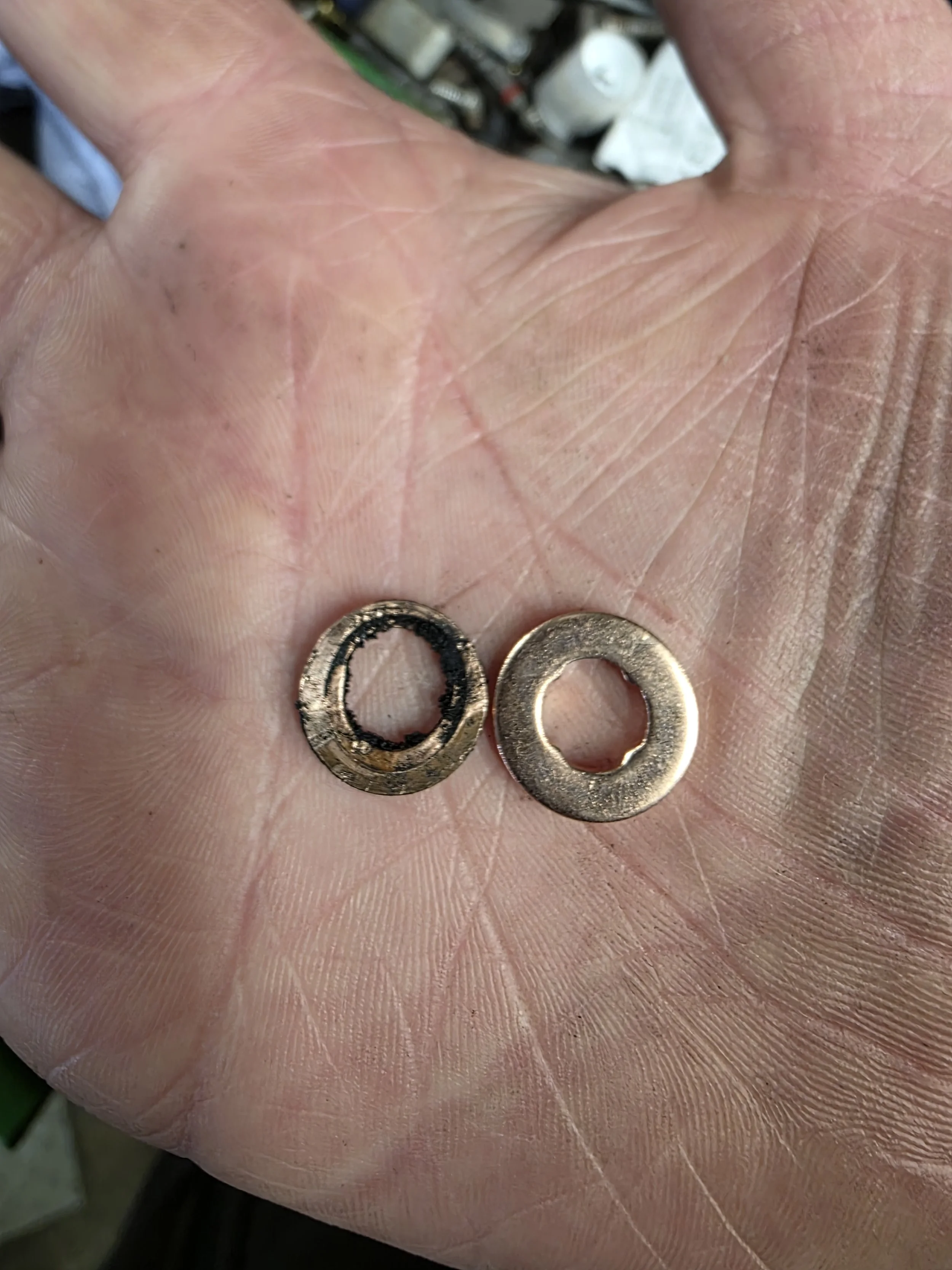 Close-up of a person's palm holding two small copper diesel injector washers, with visible fingerprints and skin texture. One failed washer and one new part