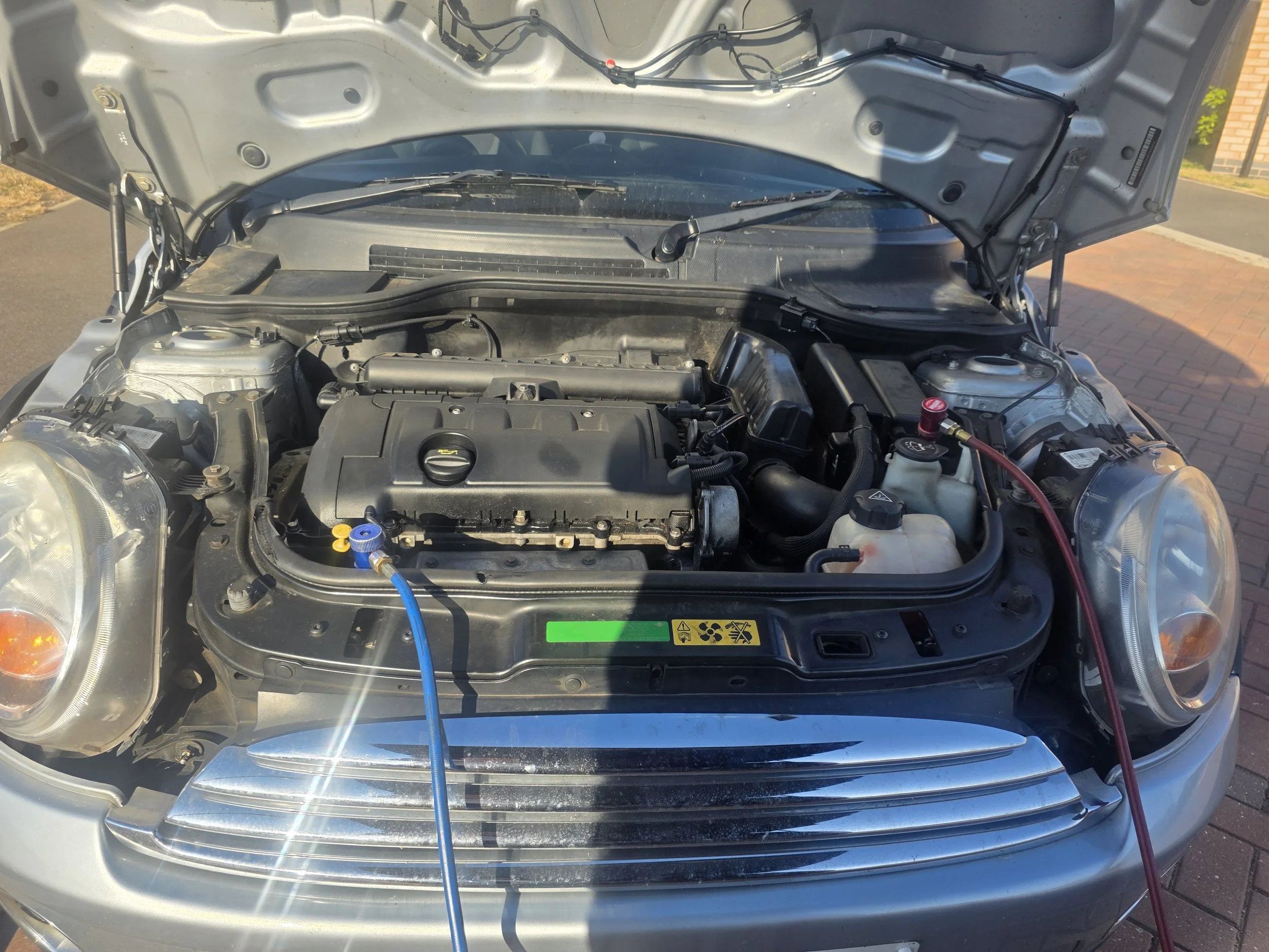 View of a car engine with the hood open, showing various engine components, wires, and hoses, parked outdoors on a sunny day. Mini air conditioning service.