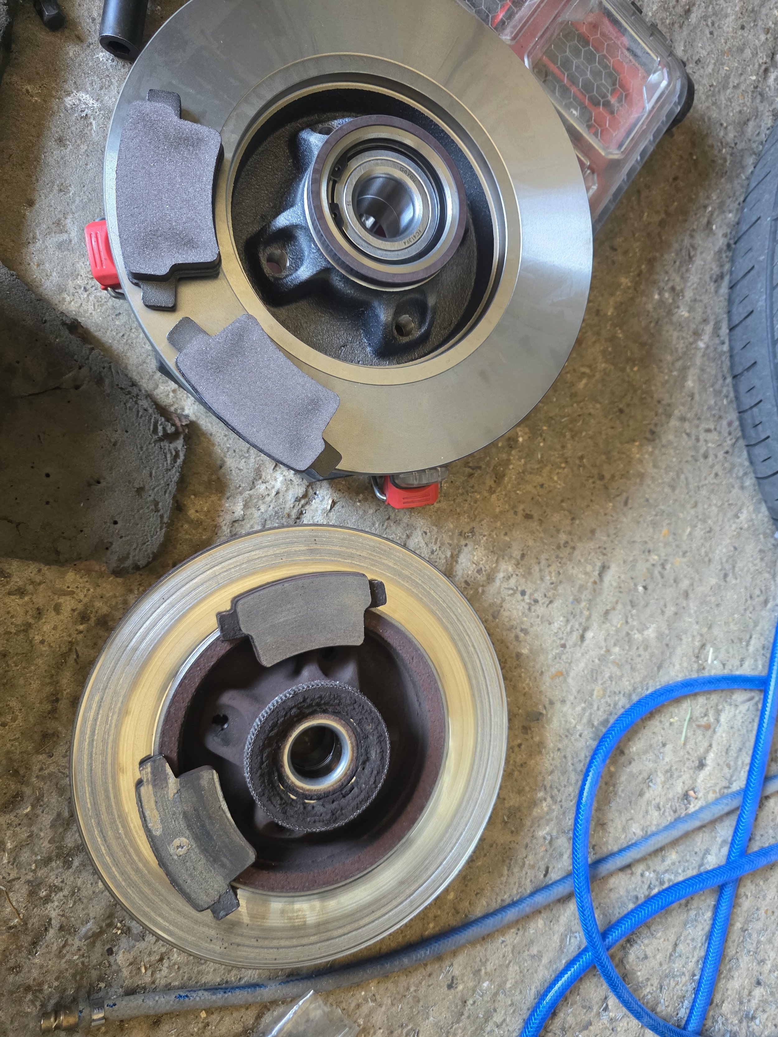 Two large automotive brake discs with brake pads and bearings, on a concrete surface with blue hoses nearby. old part and new parts