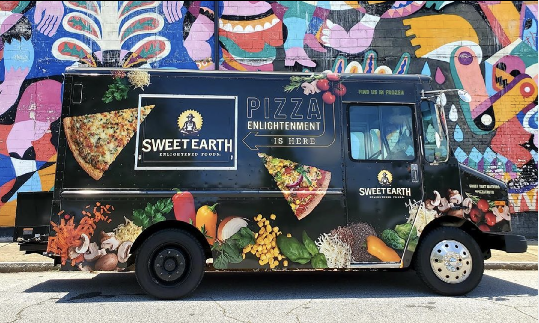 A colorful food truck with vibrant artwork and images of pizza, vegetables, and mushrooms. The truck displays a logo for 'Sweet Earth' with the slogan 'Enlightened Foods' and promotes pizza enlightenment. There are fresh ingredients, such as cherry tomatoes, peppers, onions, and greens, depicted on the lower part of the truck.