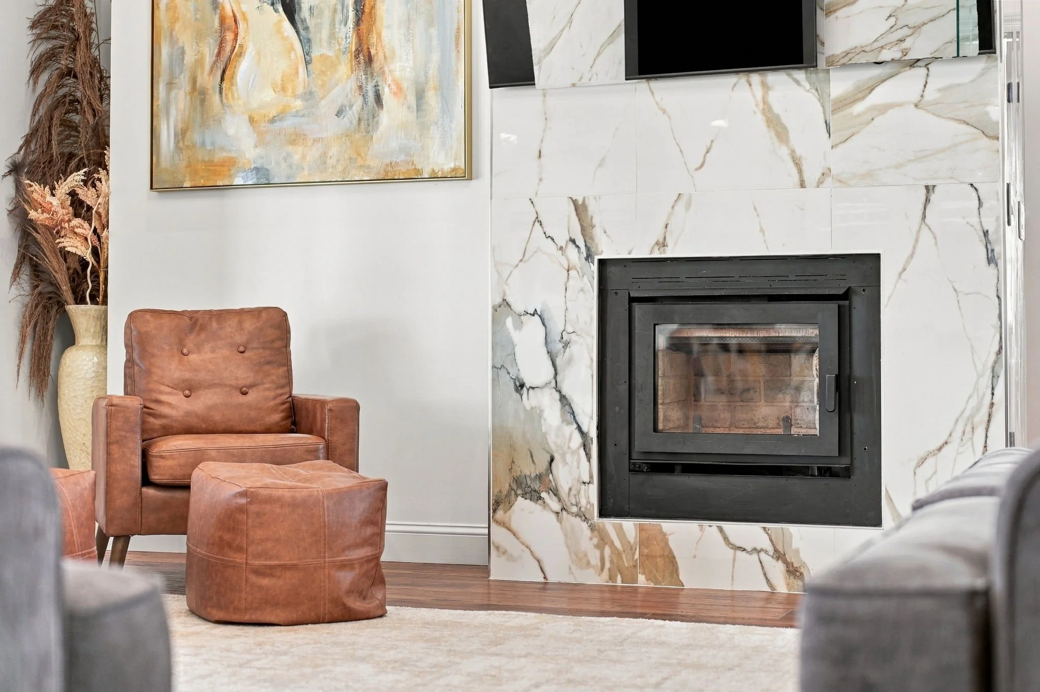 Modern living room with a marble-tile fireplace, leather armchair and ottoman, wall-mounted TV, abstract artwork, and decorative vase