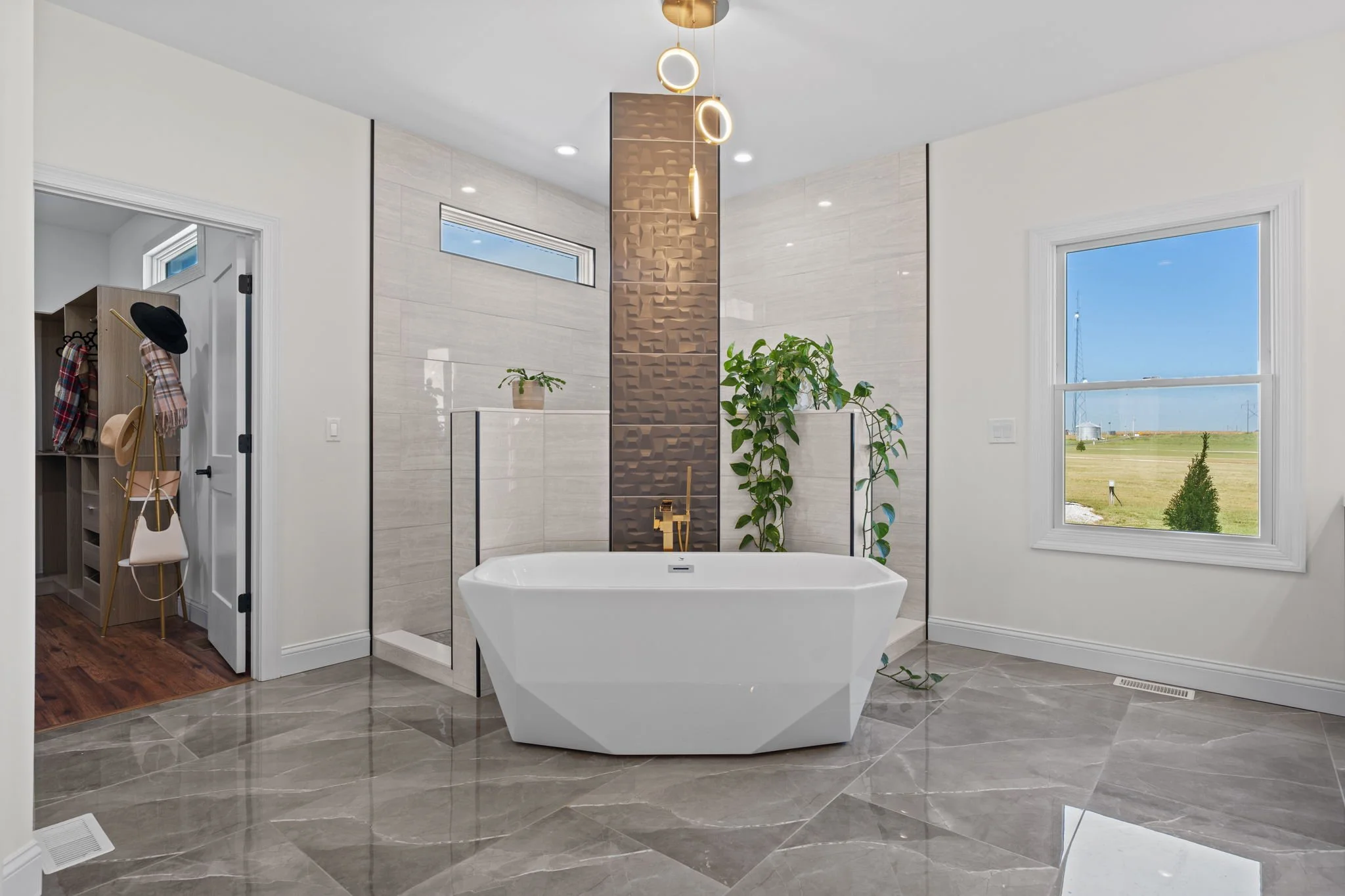 Modern luxury bathroom with freestanding bathtub, glass-enclosed shower, pendant lights, indoor plants, and large window overlooking a field