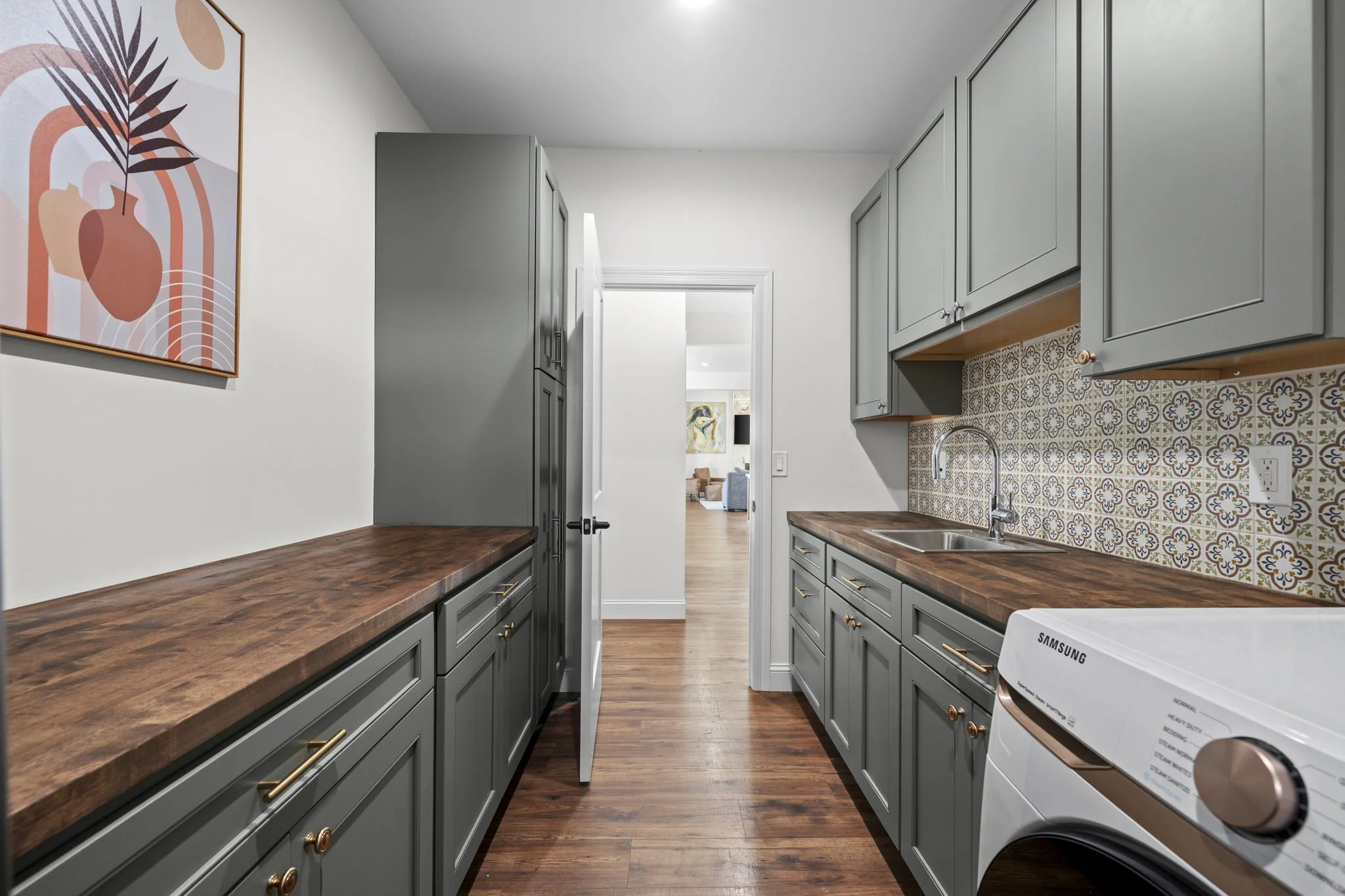 Modern laundry room with dark gray cabinets, wood countertops, patterned tile backsplash, stainless steel sink, and front-loading Samsung washer
