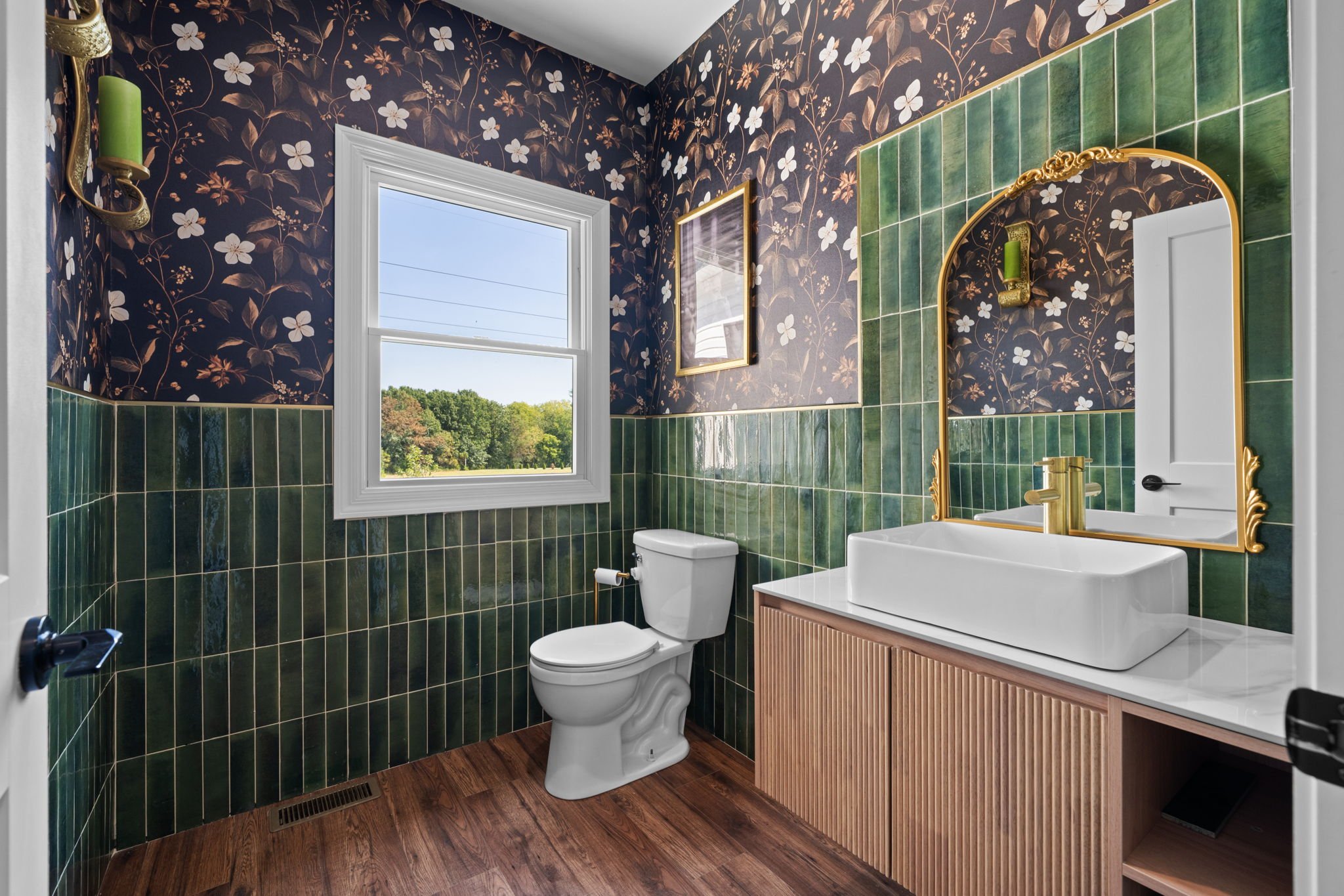Stylish powder room with dark floral wallpaper, green tile walls, a white vessel sink, gold-framed mirror, toilet, and window.