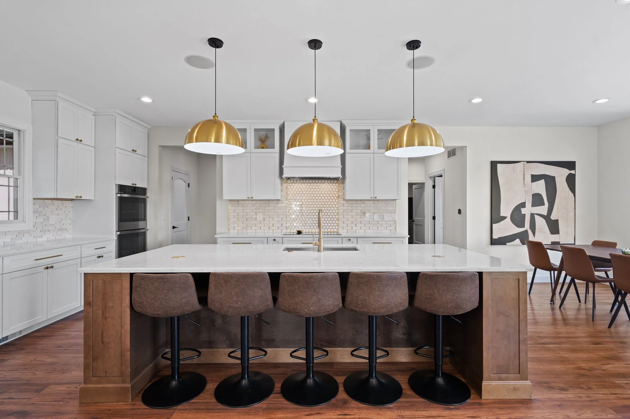 Modern white kitchen with a large marble island, five bar stools, gold pendant lights, and dining area