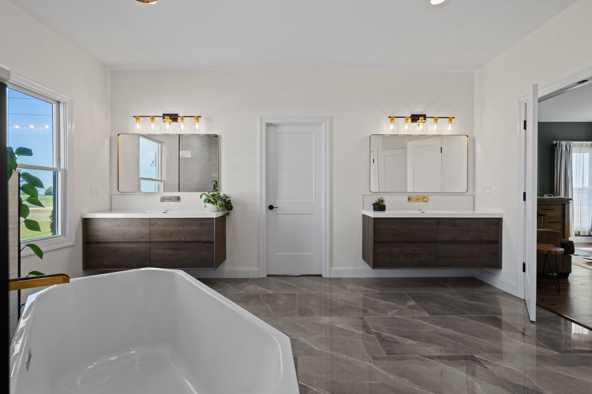 Modern bathroom with freestanding bathtub, dual dark wood vanities, gold-framed mirrors, and large windows