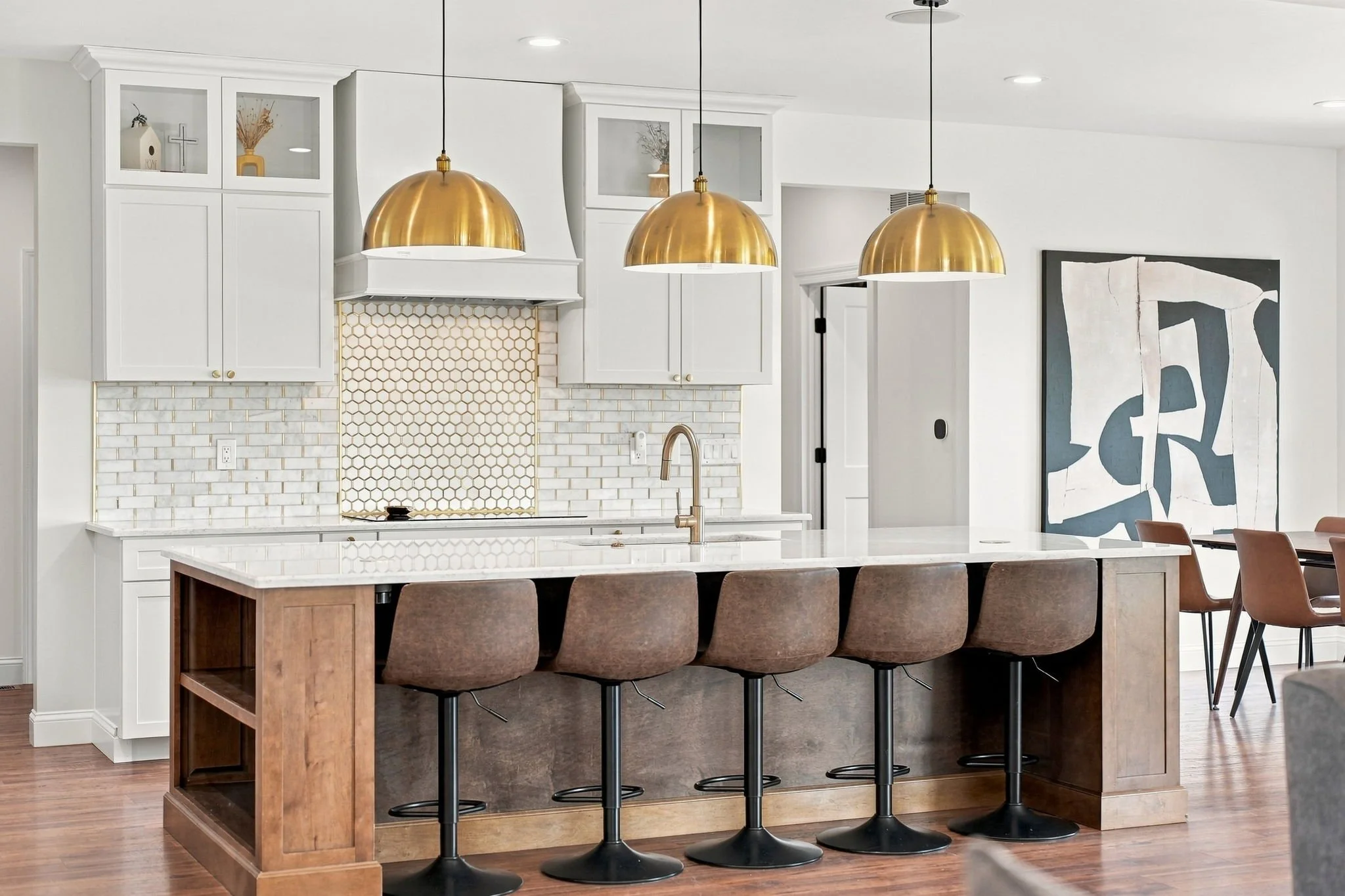 Bright modern kitchen with white cabinetry, marble island, gold pendant lights, hexagon tile backsplash, and brown bar stools
