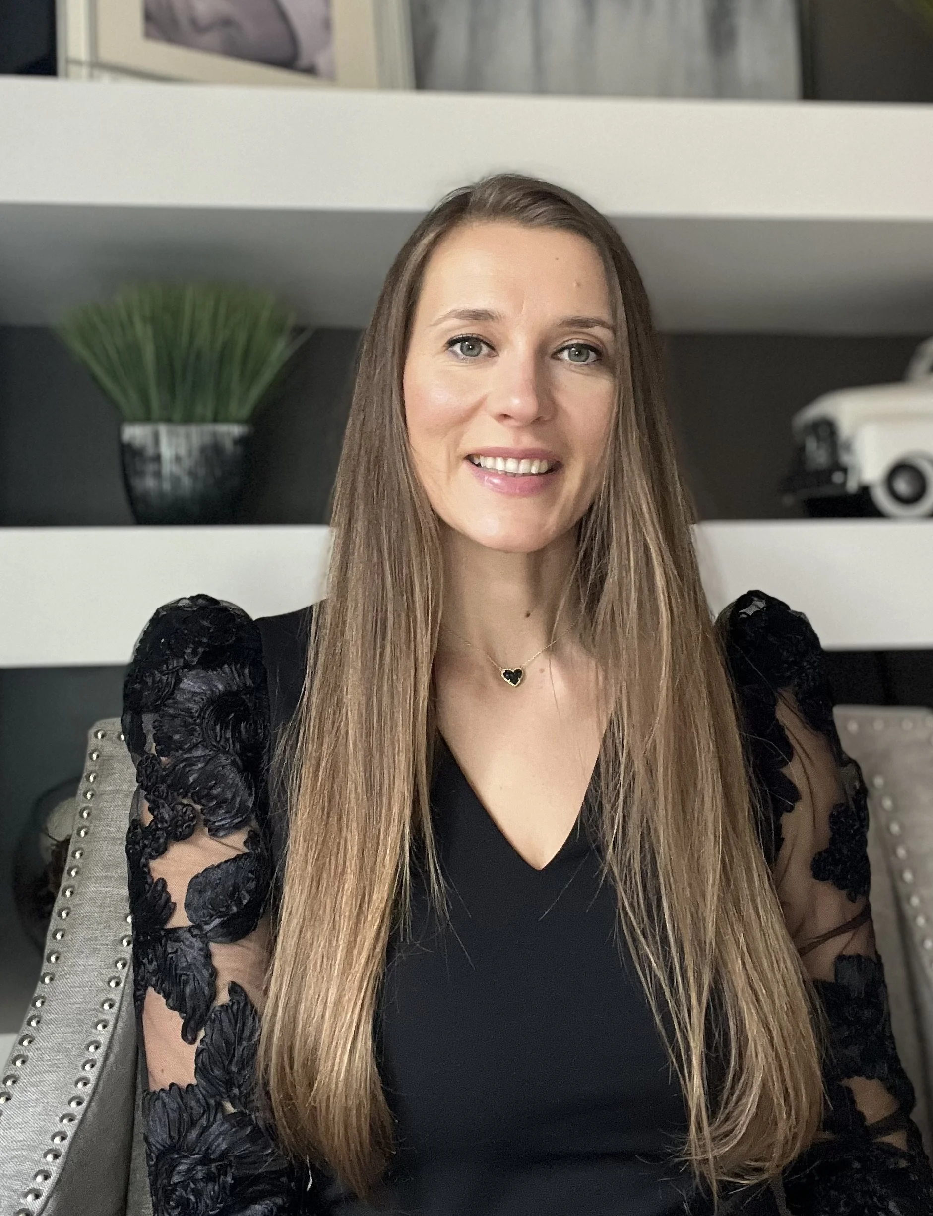 Smiling woman with long brown hair wearing a black lace-sleeved top and heart necklace, seated indoors.