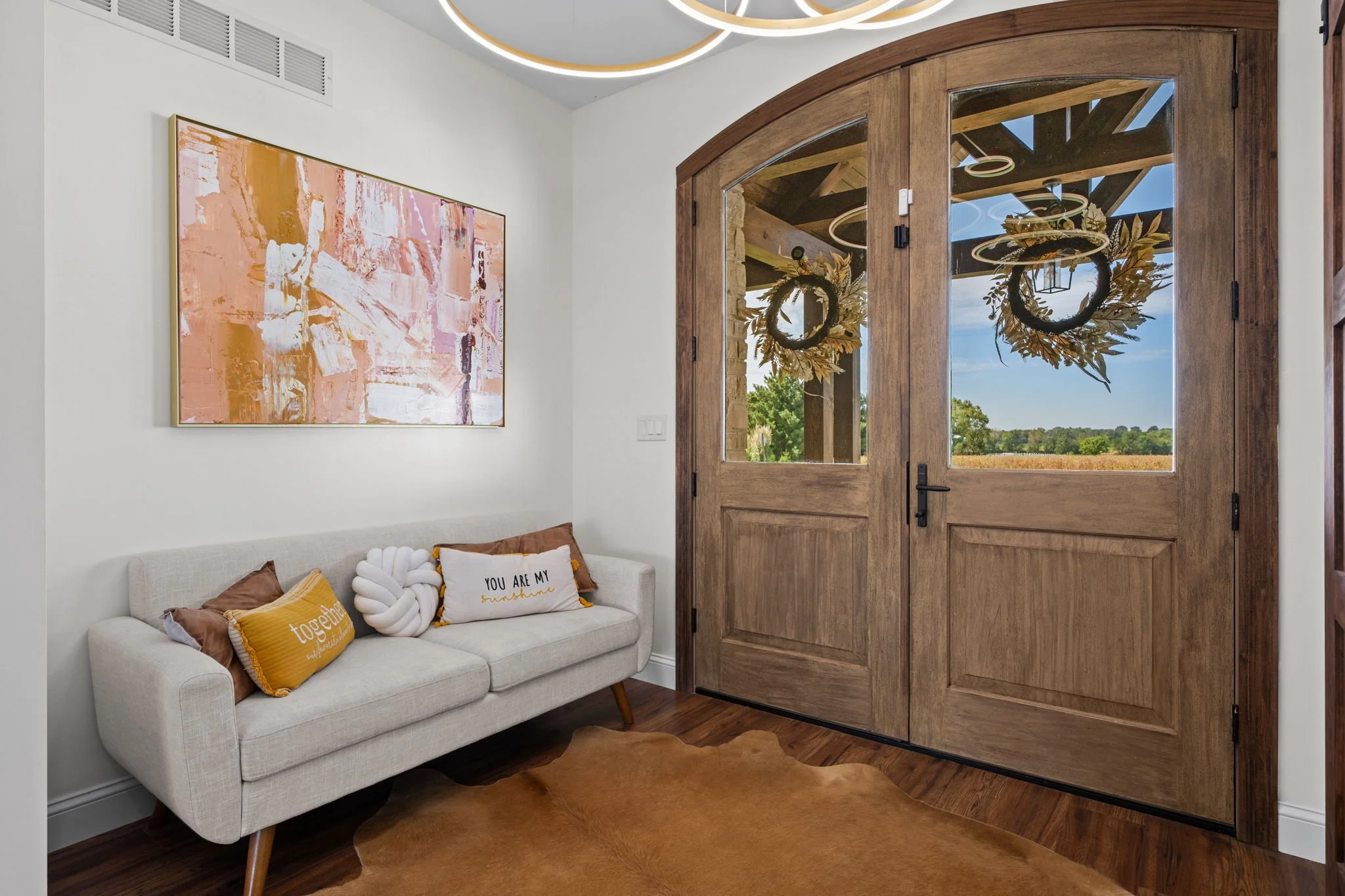 Bright entryway with a white sofa, colorful pillows, abstract wall art, wood-and-glass double doors, and a cowhide rug.