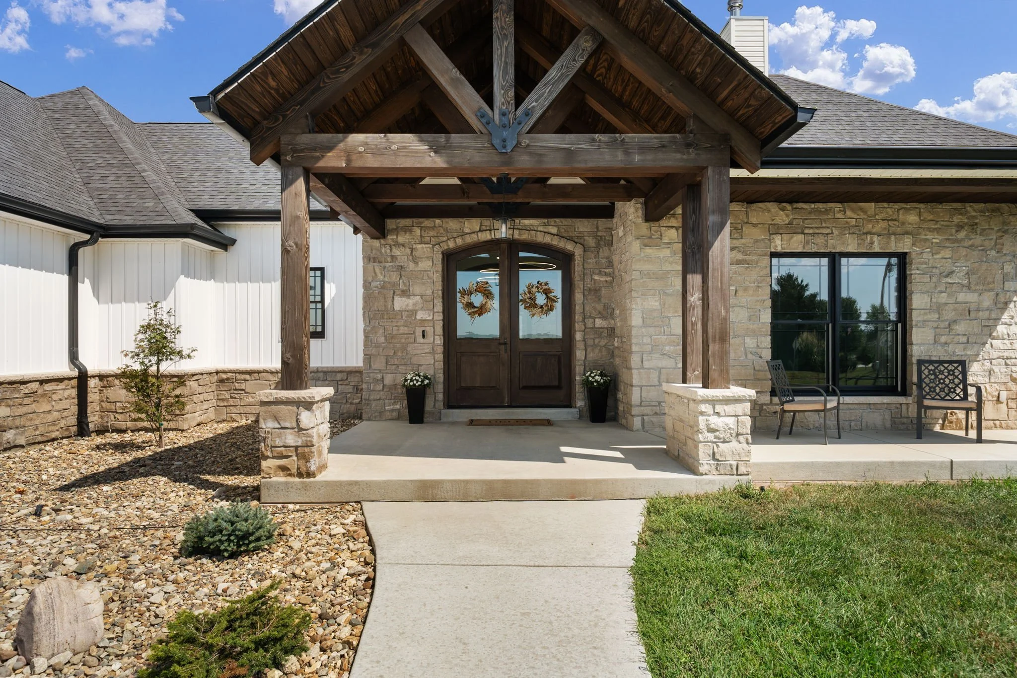 Stone-front home entrance with a large timber portico, double wooden doors, rock landscaping, and outdoor chairs.