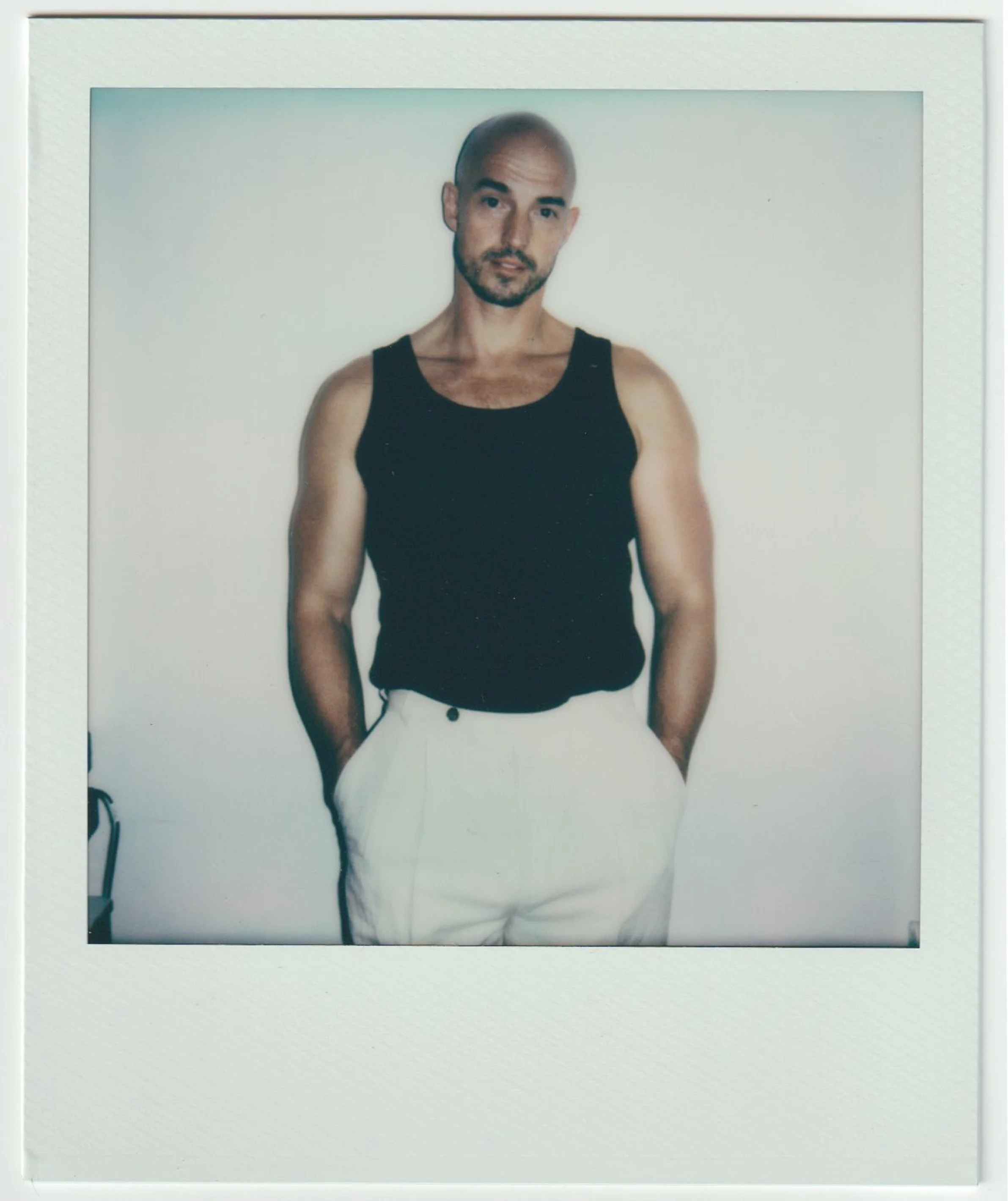 A man with a shaved head, beard, and mustache, wearing a black tank top, standing against a plain white background.