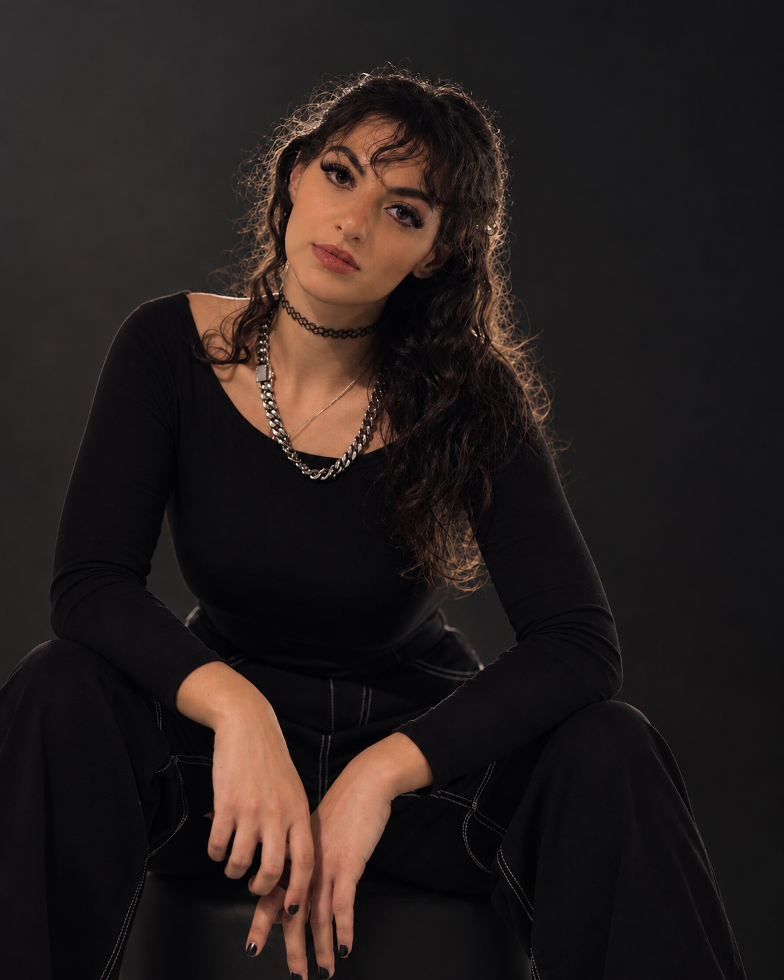 A young woman with long, curly hair wearing a black long-sleeve shirt, layered necklaces, and a choker, sitting against a dark background, looking at the camera.