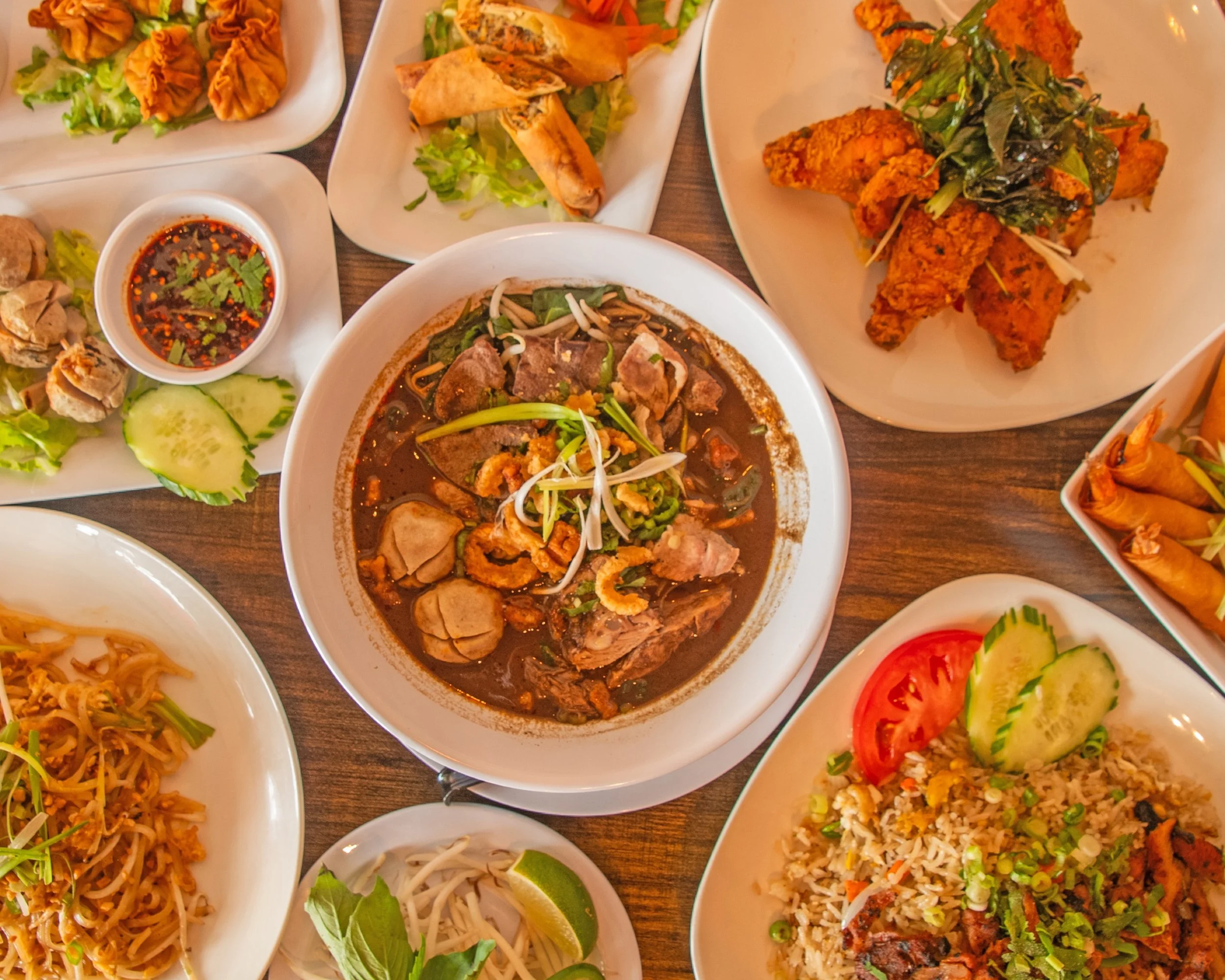 A variety of Asian dishes on white plates, including fried chicken with herbs, spring rolls, noodle stir-fry, and bowls of soup, with small bowls of soy sauce and sliced cucumbers and tomatoes.