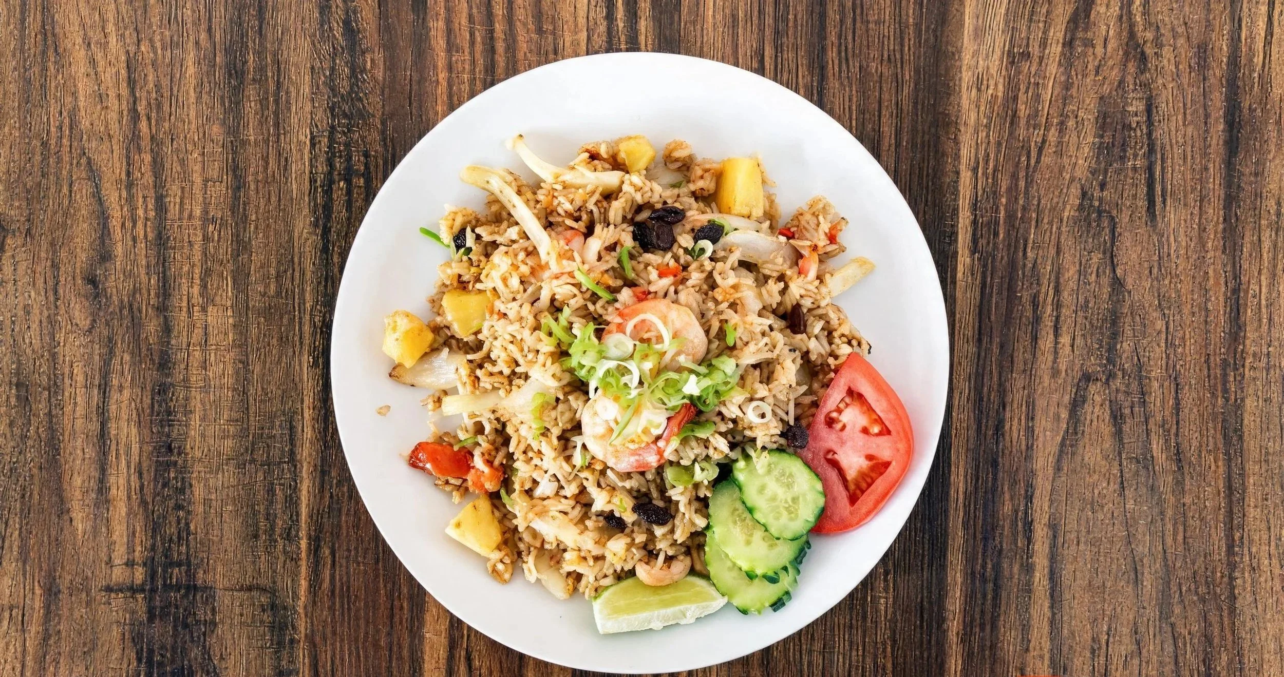 A white plate of fried rice with vegetables and shrimp, garnished with sliced cucumber, tomato, green onions, and pineapple on a rustic wooden table.