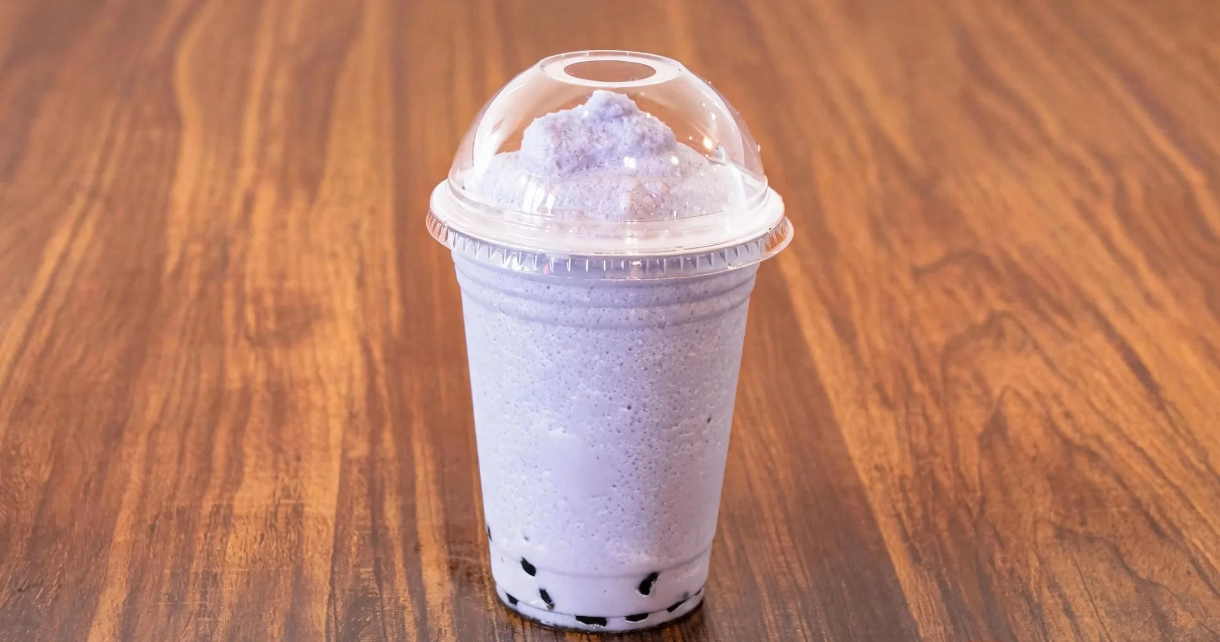 A tall transparent plastic cup filled with a light purple bubblegum-flavored milk tea topped with purple whipped cream and black tapioca pearls at the bottom, placed on a wooden table.