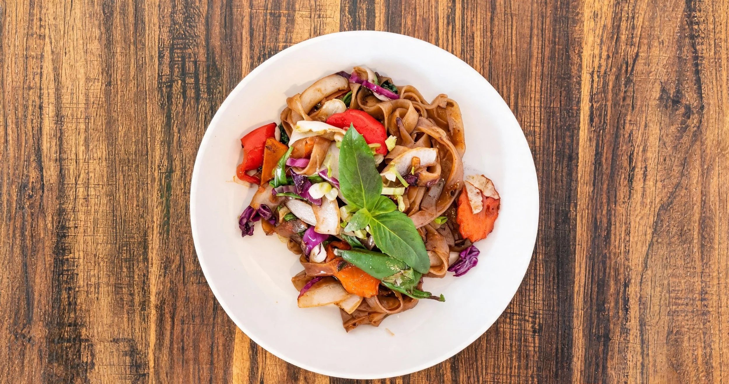 A white plate with stir-fried noodles and colorful vegetables, garnished with fresh green herbs, on a wooden table.