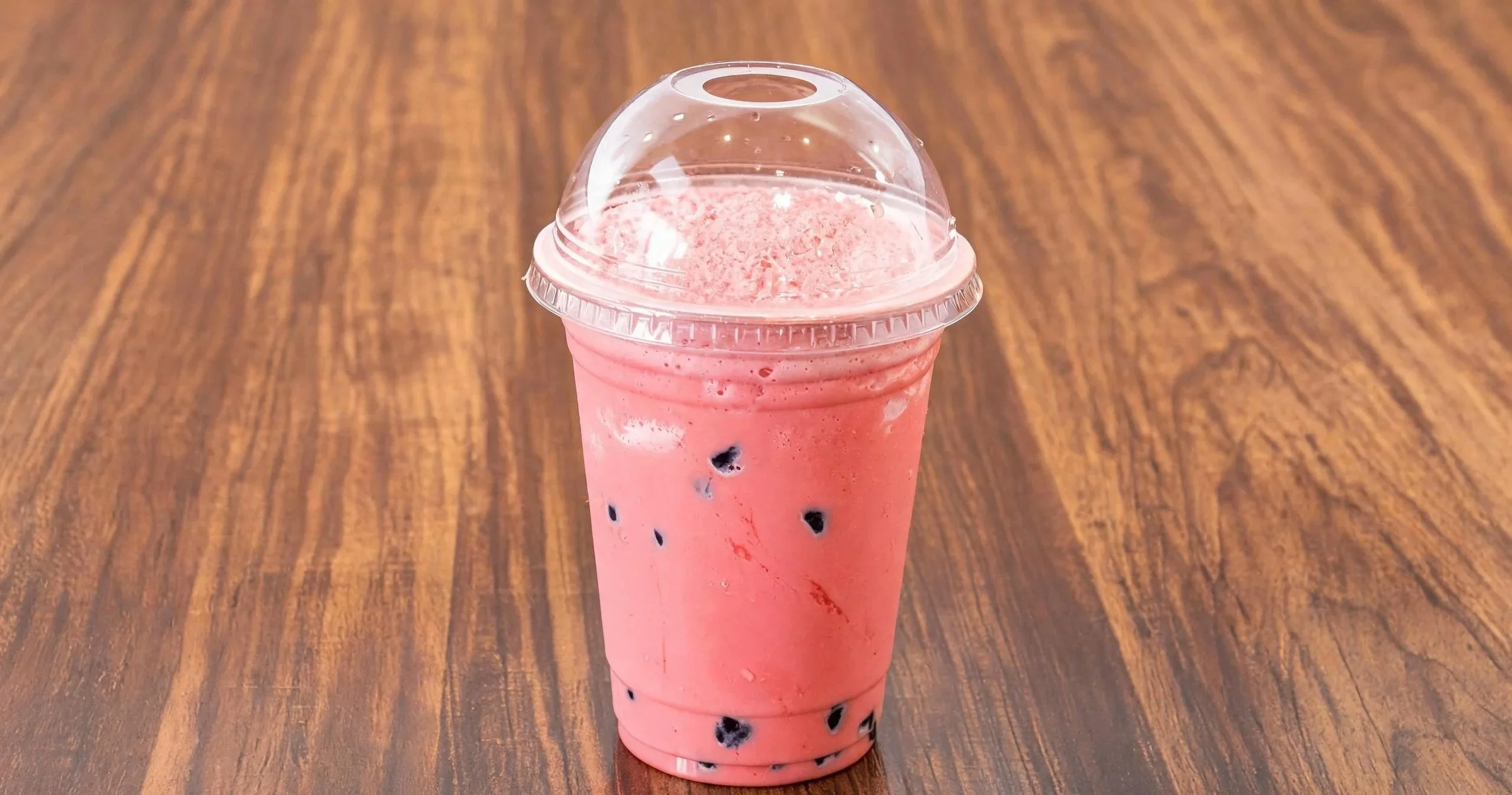 A plastic cup filled with pink strawberry-flavored bubble tea with black tapioca pearls at the bottom, placed on a wooden surface.