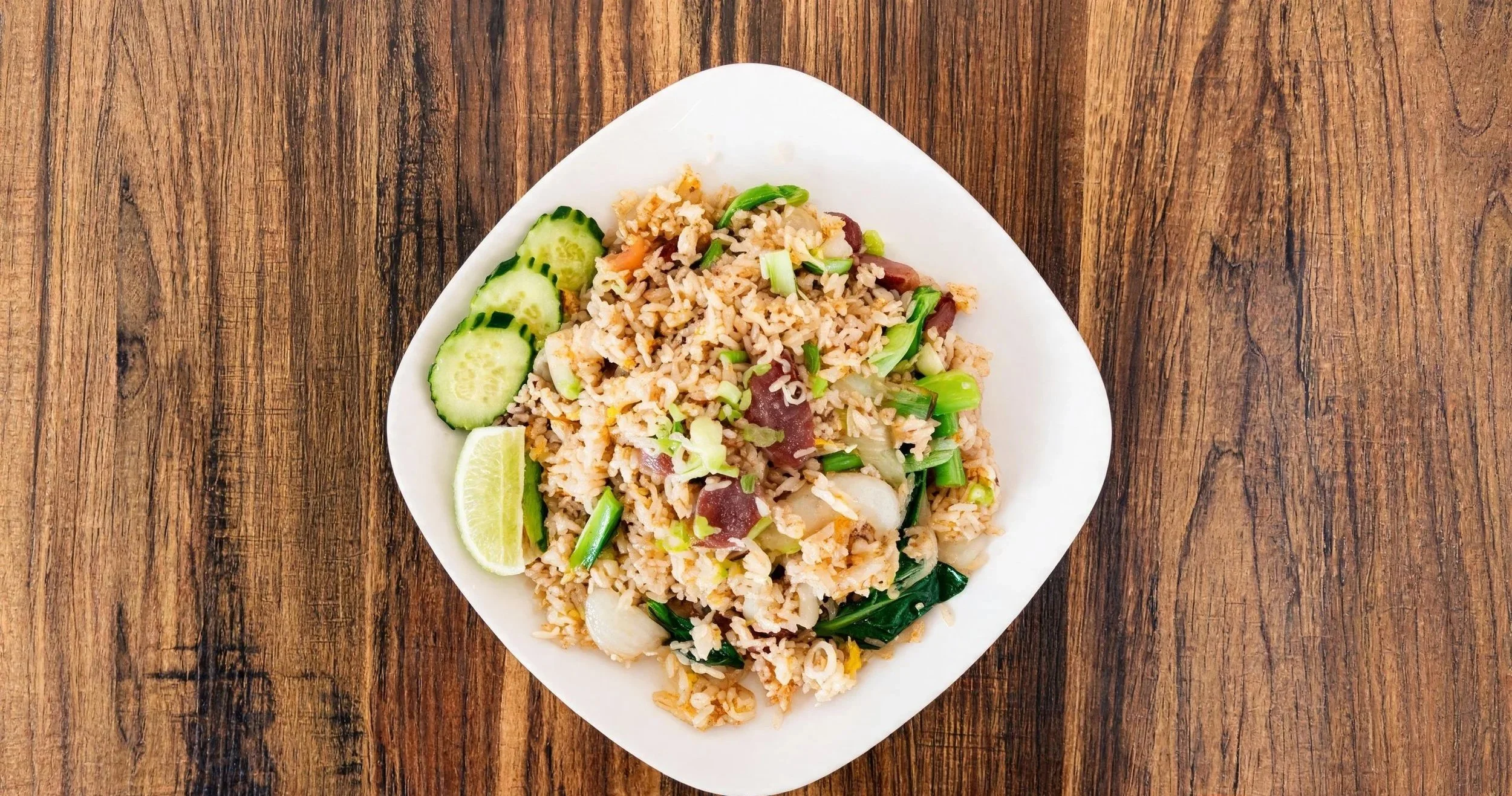 A white plate of fried rice with green onions, cucumber slices, and a lime wedge on a wooden table.