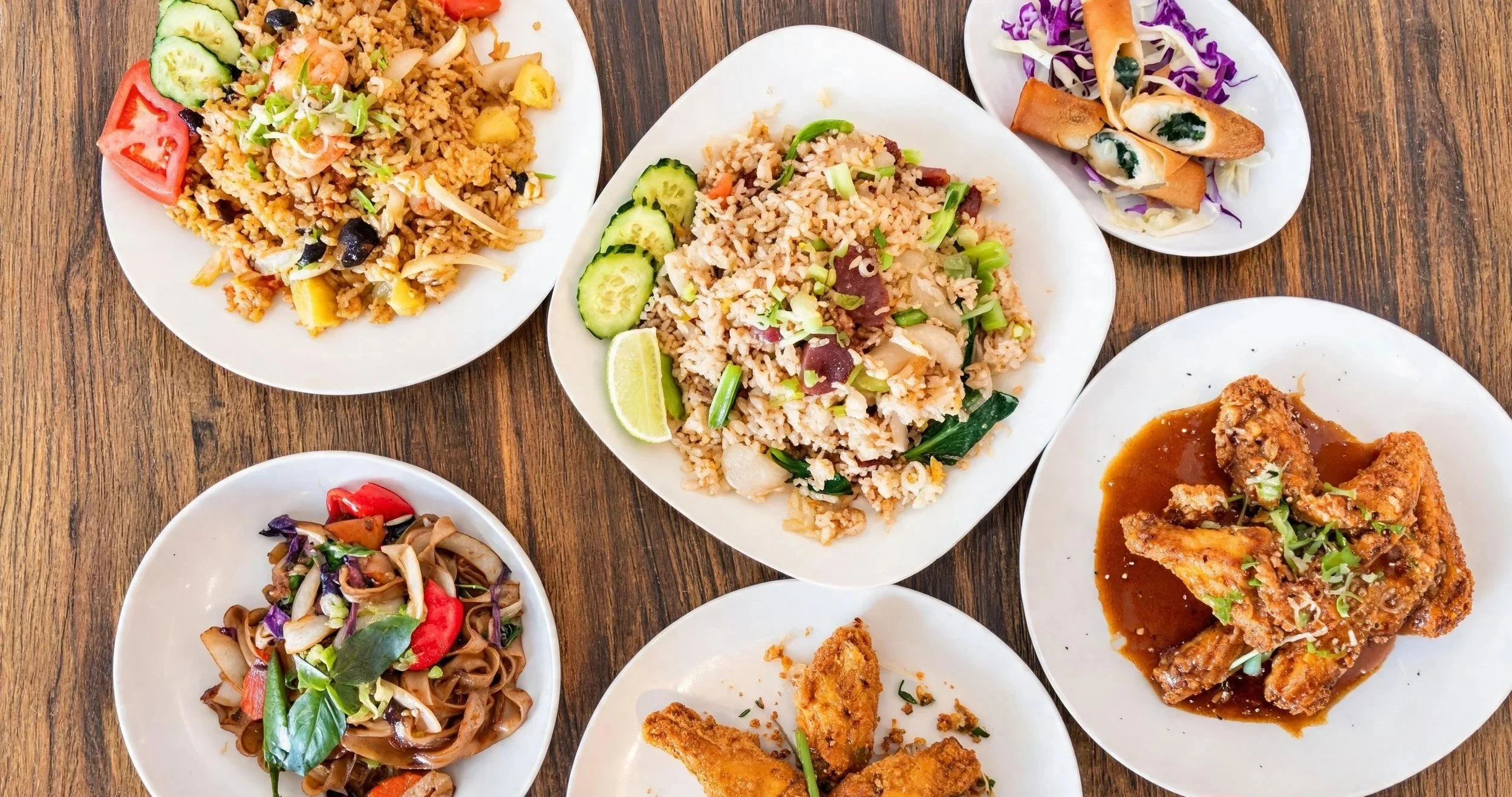 Various Asian dishes on white plates, including fried rice with cucumber and lime, stir-fried noodles with vegetables, spring rolls with purple cabbage, and fried chicken in sauce on a wooden table.