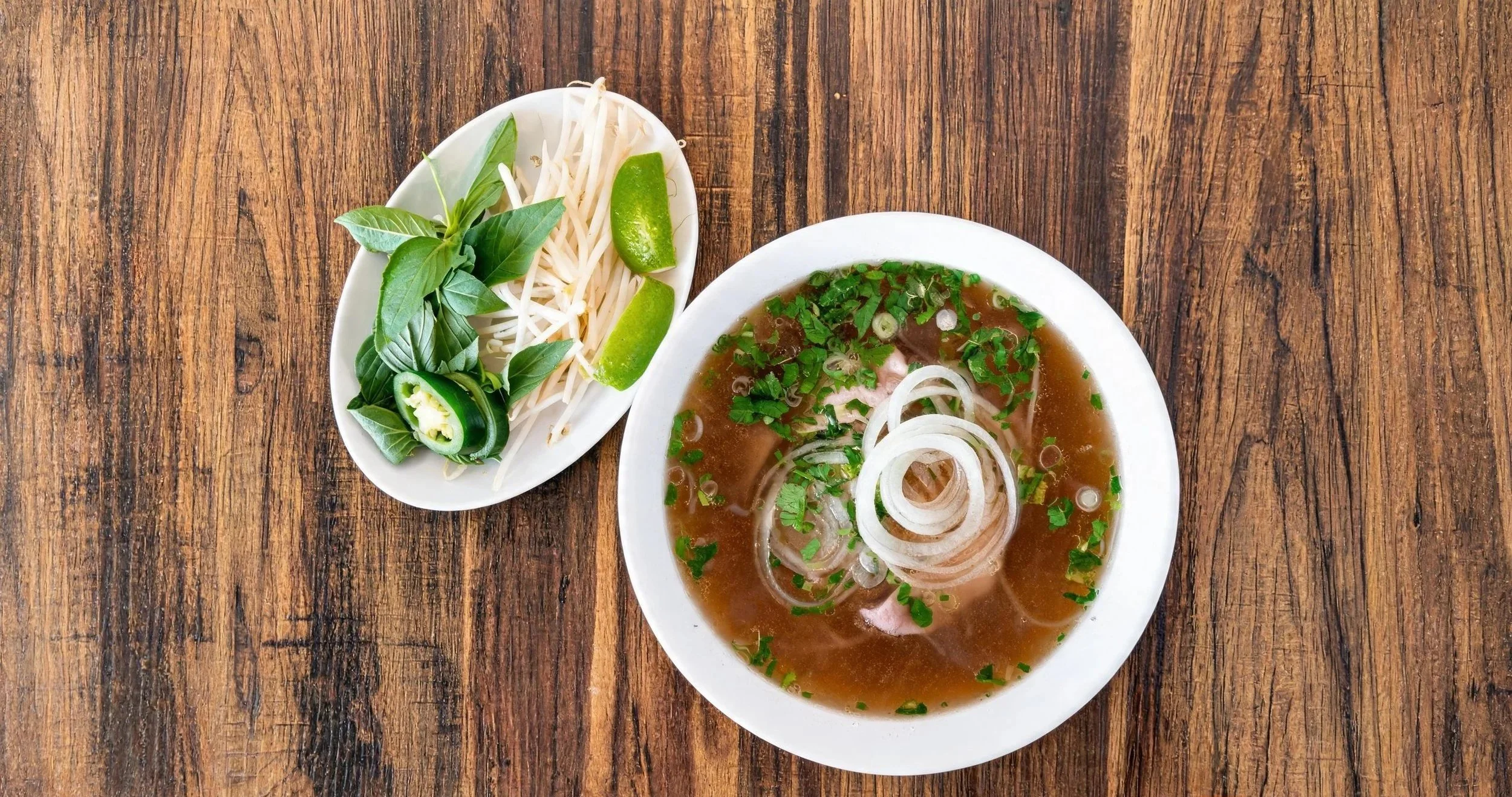 Bowl of Vietnamese pho with beef broth, sliced onions, and chopped herbs, alongside a small plate of bean sprouts, lime wedges, basil, and sliced chili pepper on a wooden table.