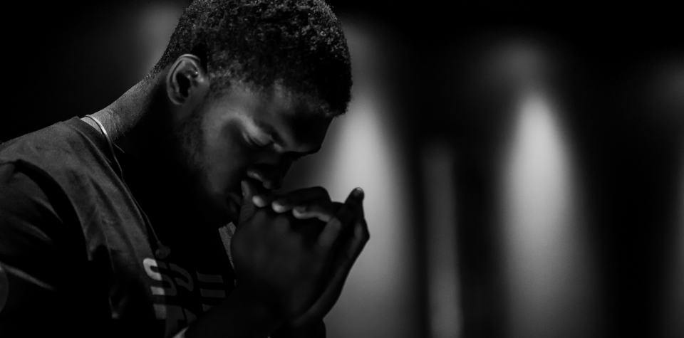 A young man praying with clasped hands in a dimly lit room.