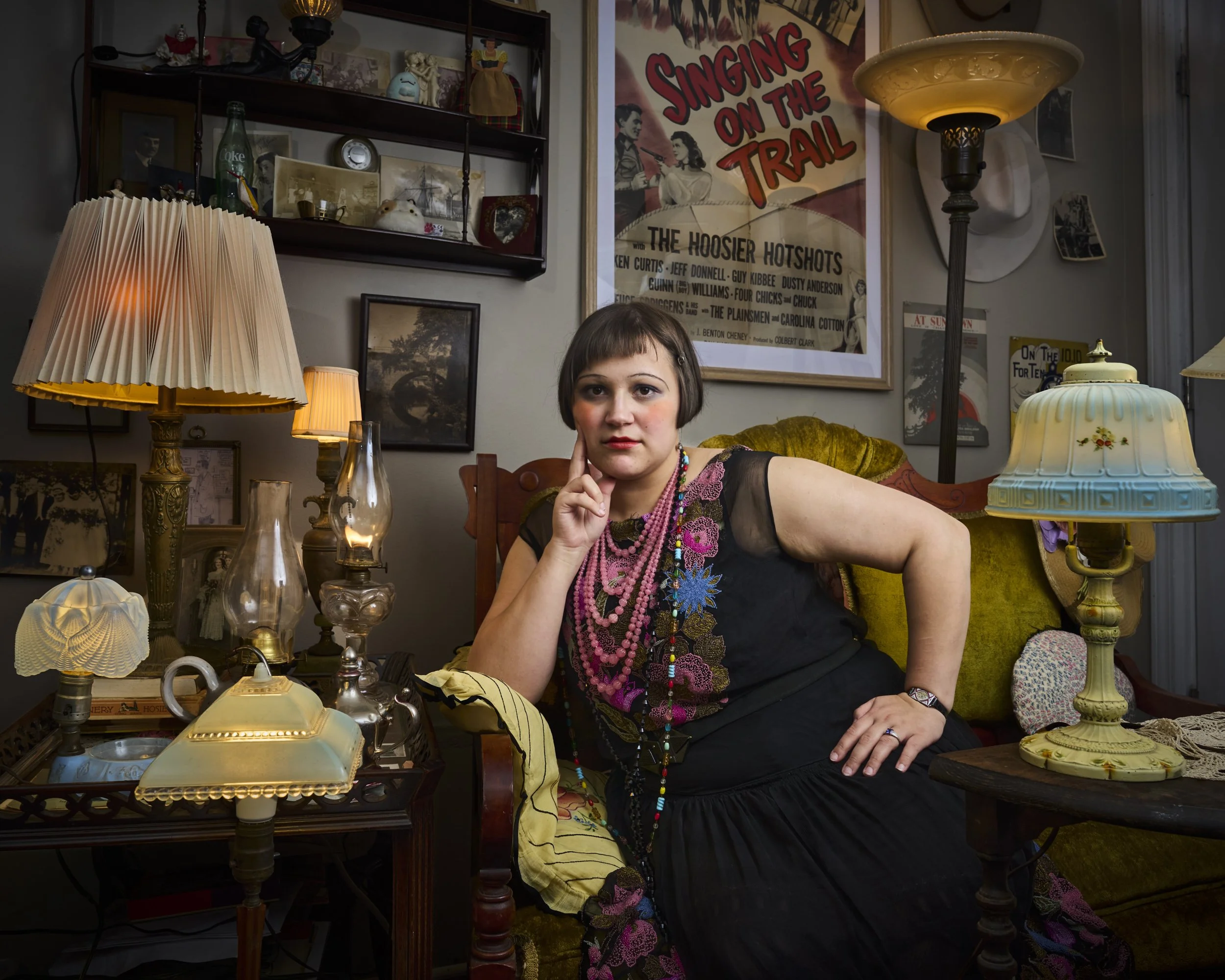 A young woman with short brown hair, wearing colorful necklaces and a black dress, sits at a cluttered table in a vintage-style room filled with lamps, framed photos, and nostalgic decorations. She rests her chin on her hand, looking at the camera. B